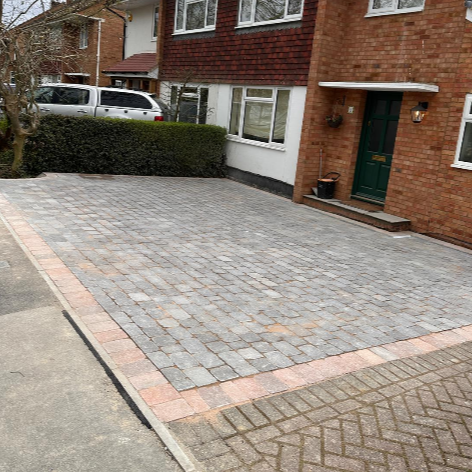 Residential driveway made of gray and red paving stones, leading to a green front door of a brick house with a large window and porch lights.