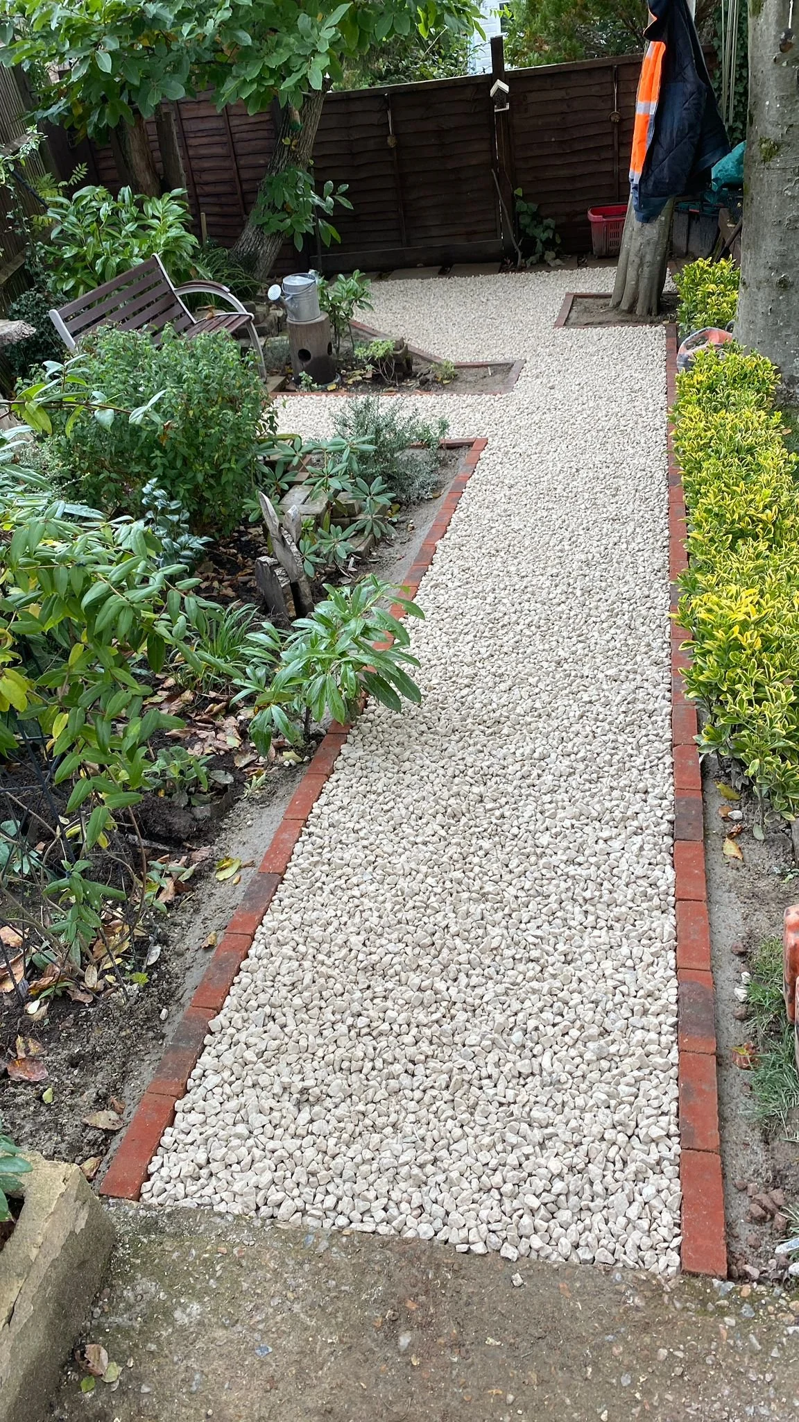 A garden pathway made of small white gravel, bordered by red bricks, with various plants and shrubs on both sides. In the background, there is a wooden fence, a bench, and some gardening tools.