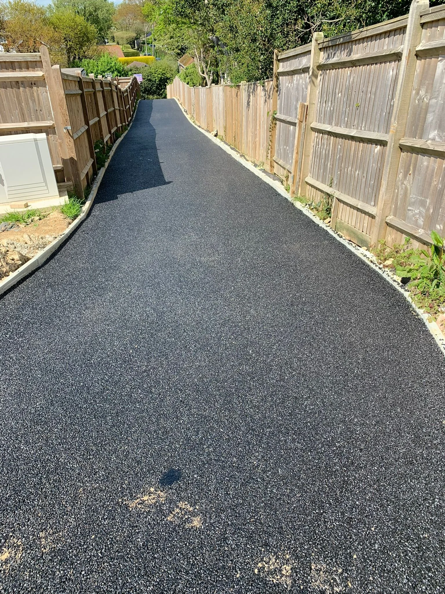A newly paved black asphalt pathway in a backyard, bordered by a wooden fence on both sides, with green trees and houses in the background.