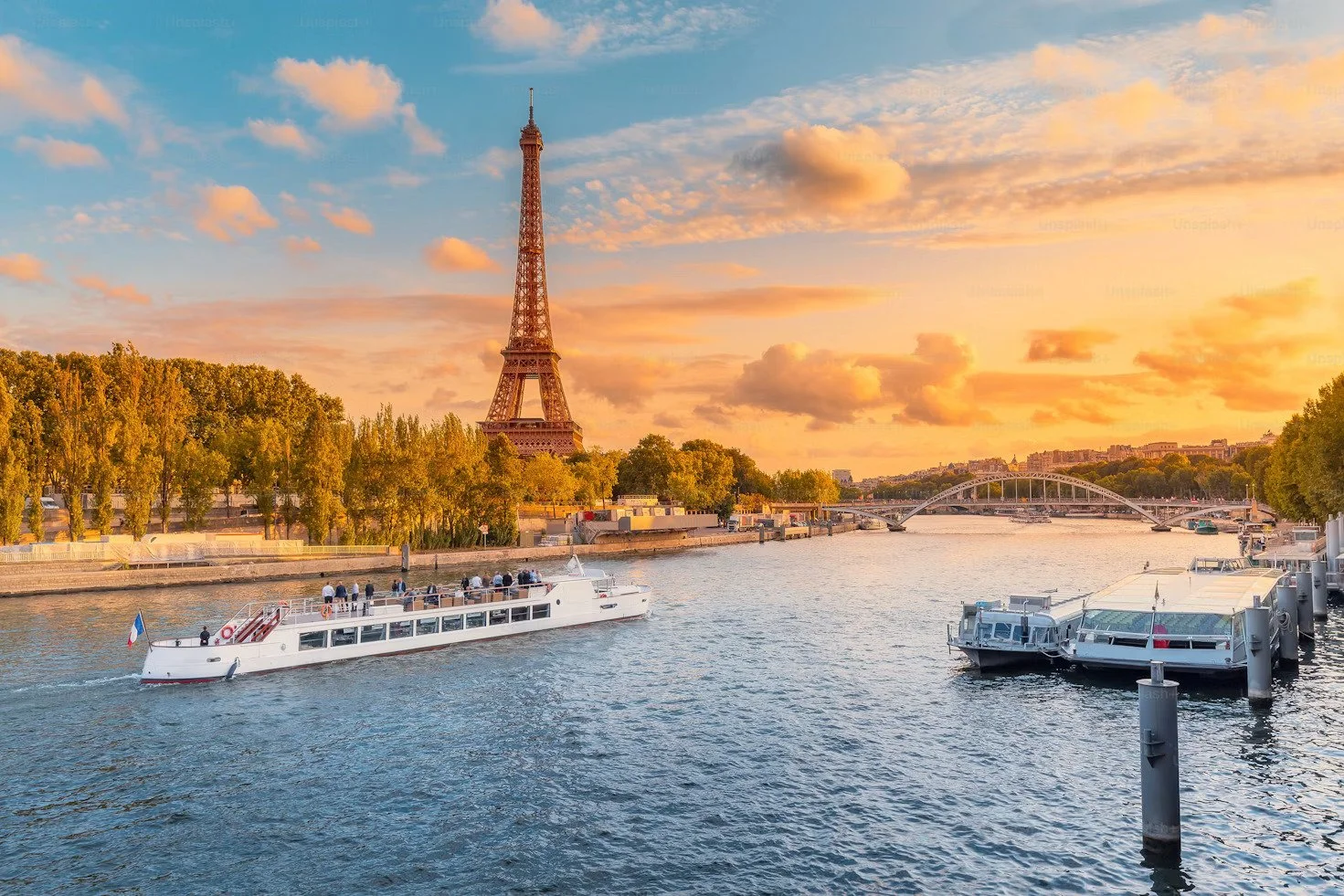 Eiffel Tower along the Seine River in Paris during sunset with boats on the water and a bridge in the background.