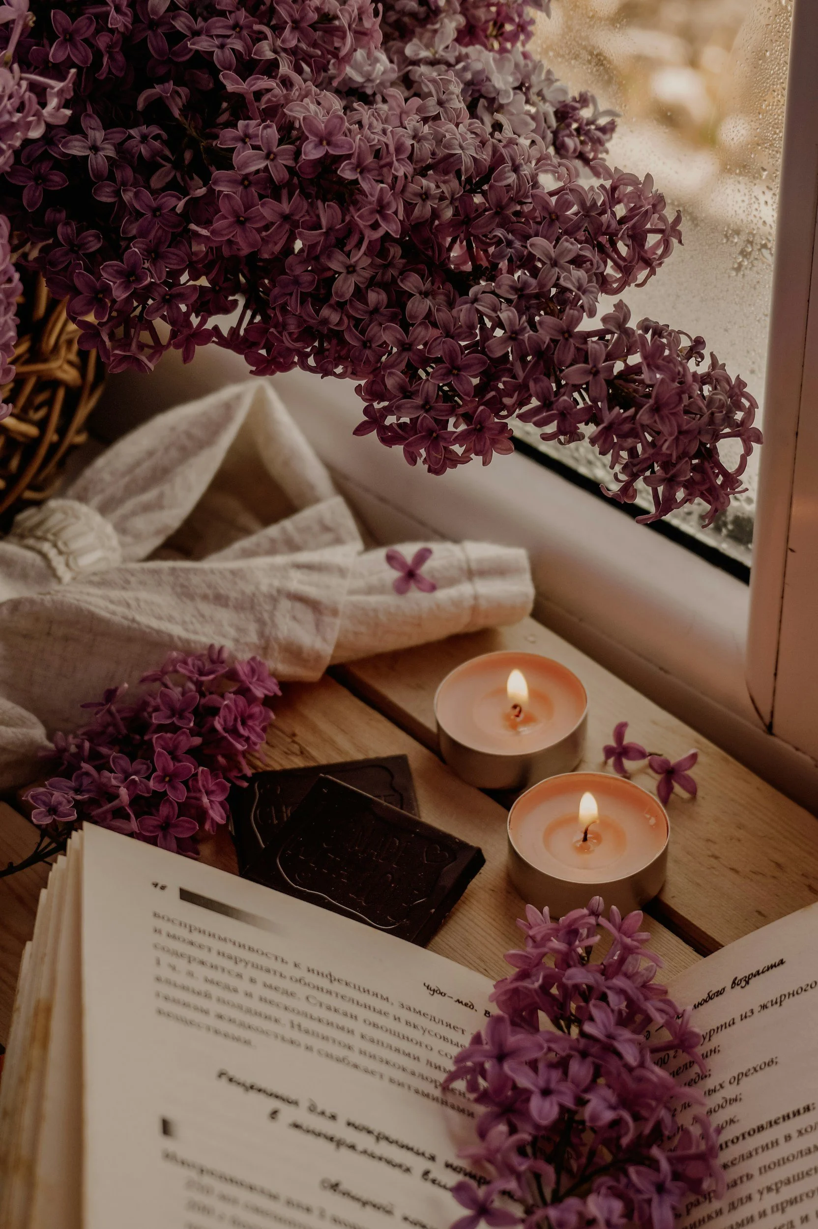 A cozy scene by a window featuring purple lilac flowers, two lit candles, an open book, and a white cloth, with raindrops visible on the window.
