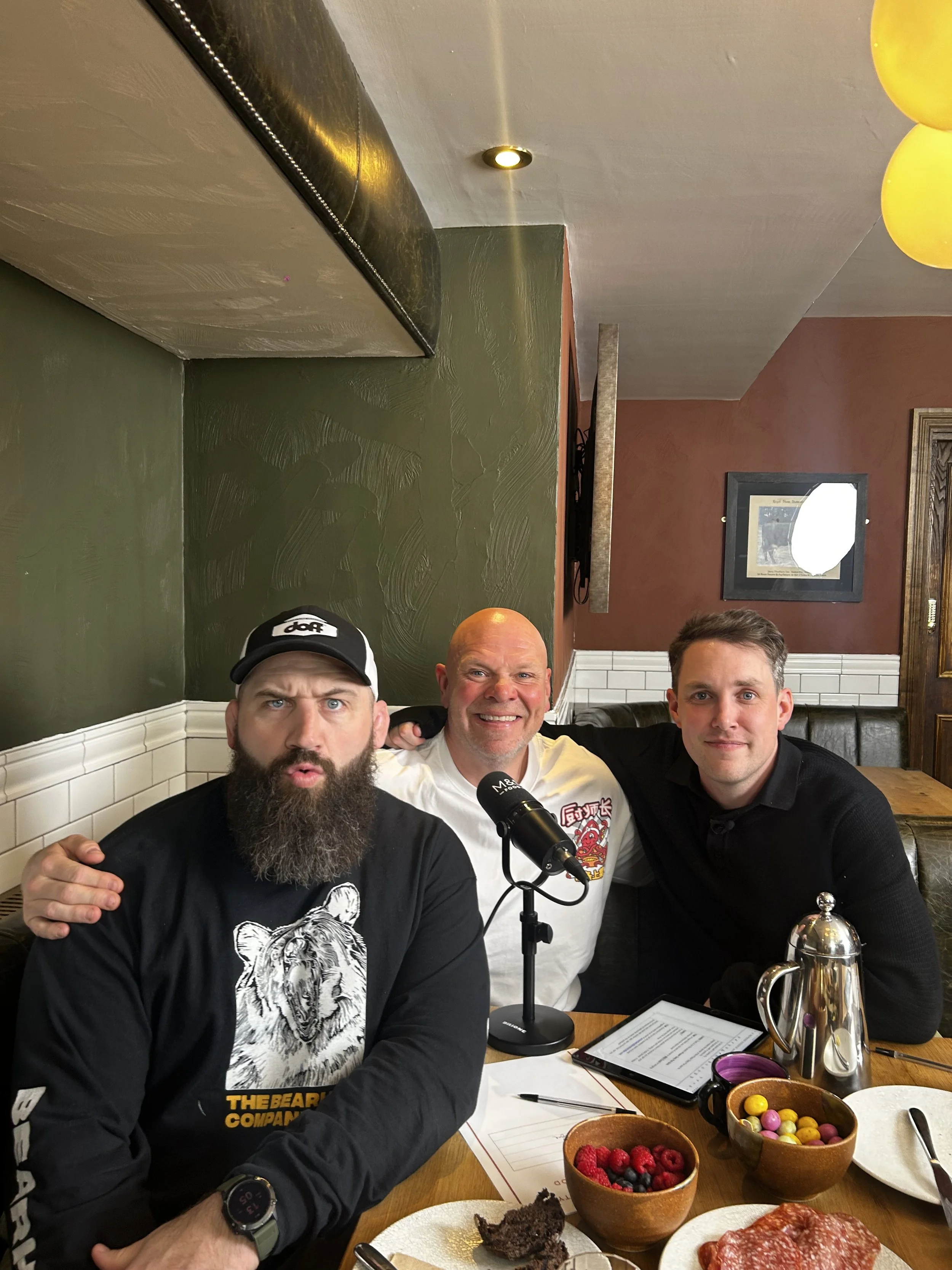 Three men sitting at a restaurant table with food and drinks, two of whom are smiling and one with a serious expression. The table has a bowl of mixed berries, a bowl of colorful candies, a slice of chocolate cake, slices of cured meat, and a tablet device. One man is wearing a black hoodie and cap, another is bald and smiling, and the third is a young man in a black shirt.