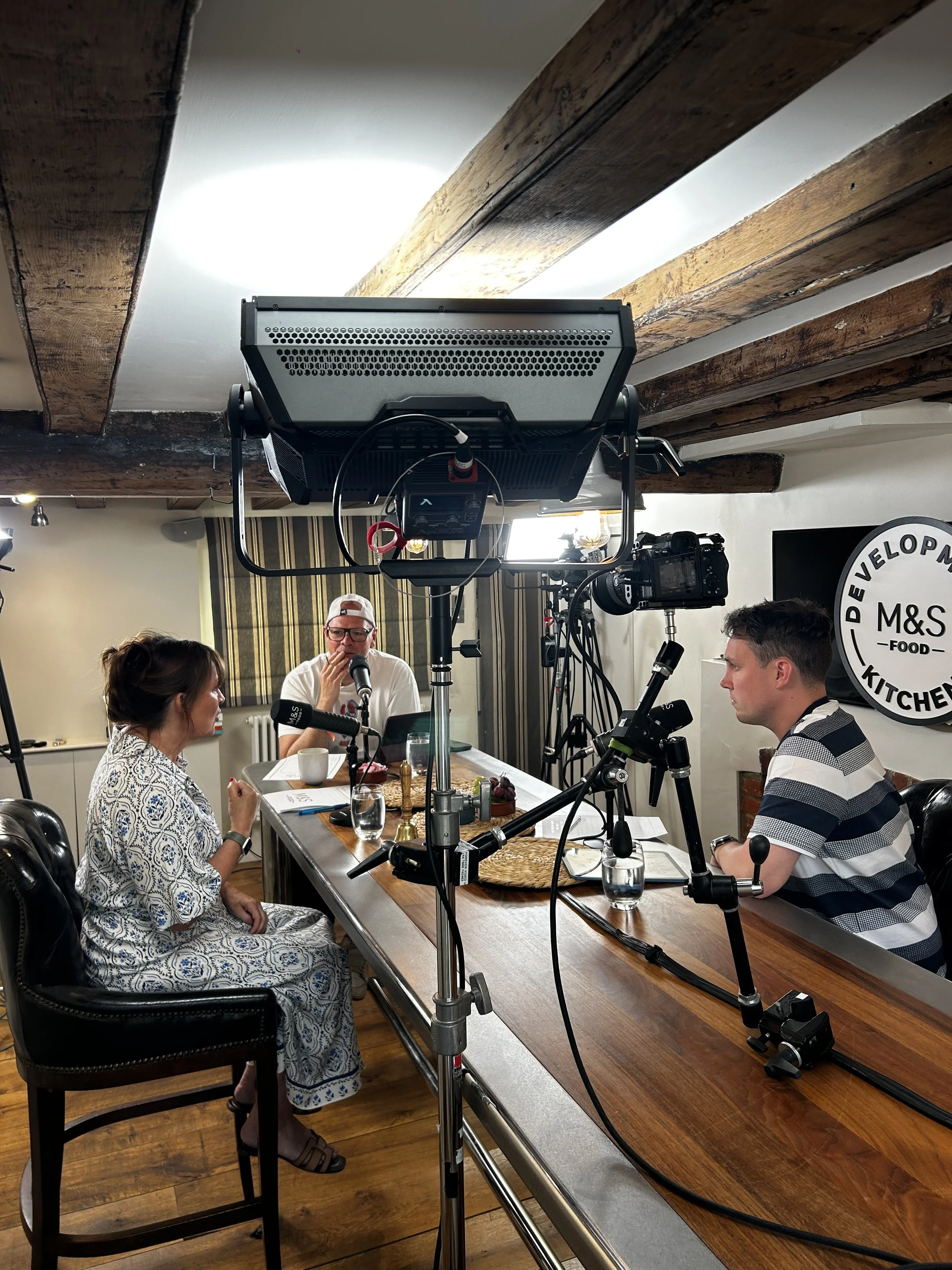 People seated at a dining table having a discussion or interview, with recording and filming equipment around them.