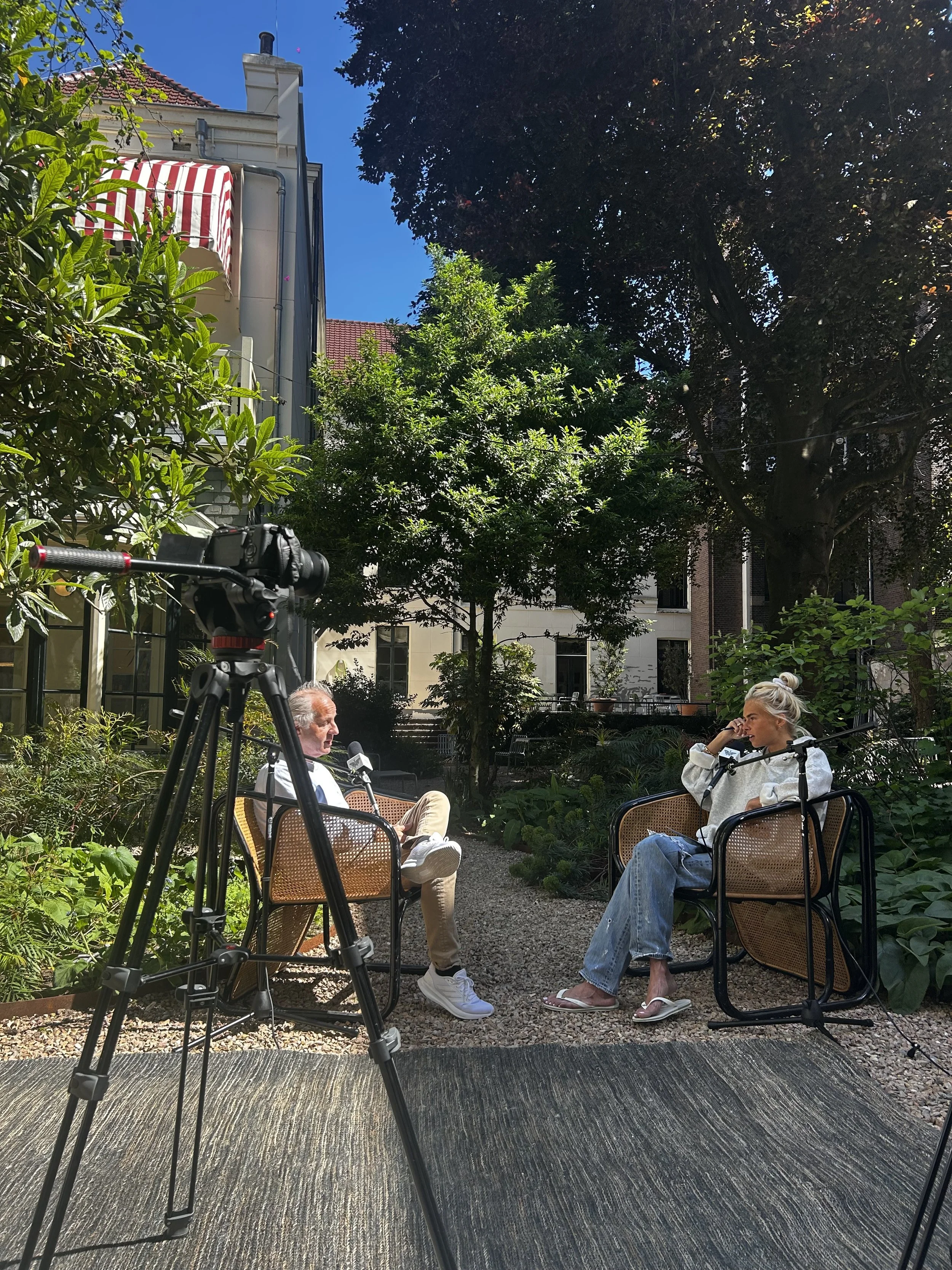 Two people are sitting outdoors in a garden, engaged in a conversation. A camera on a tripod is set up in front of them capturing the interview or discussion. The scene is surrounded by lush green trees and plants, with buildings in the background and a clear blue sky overhead.