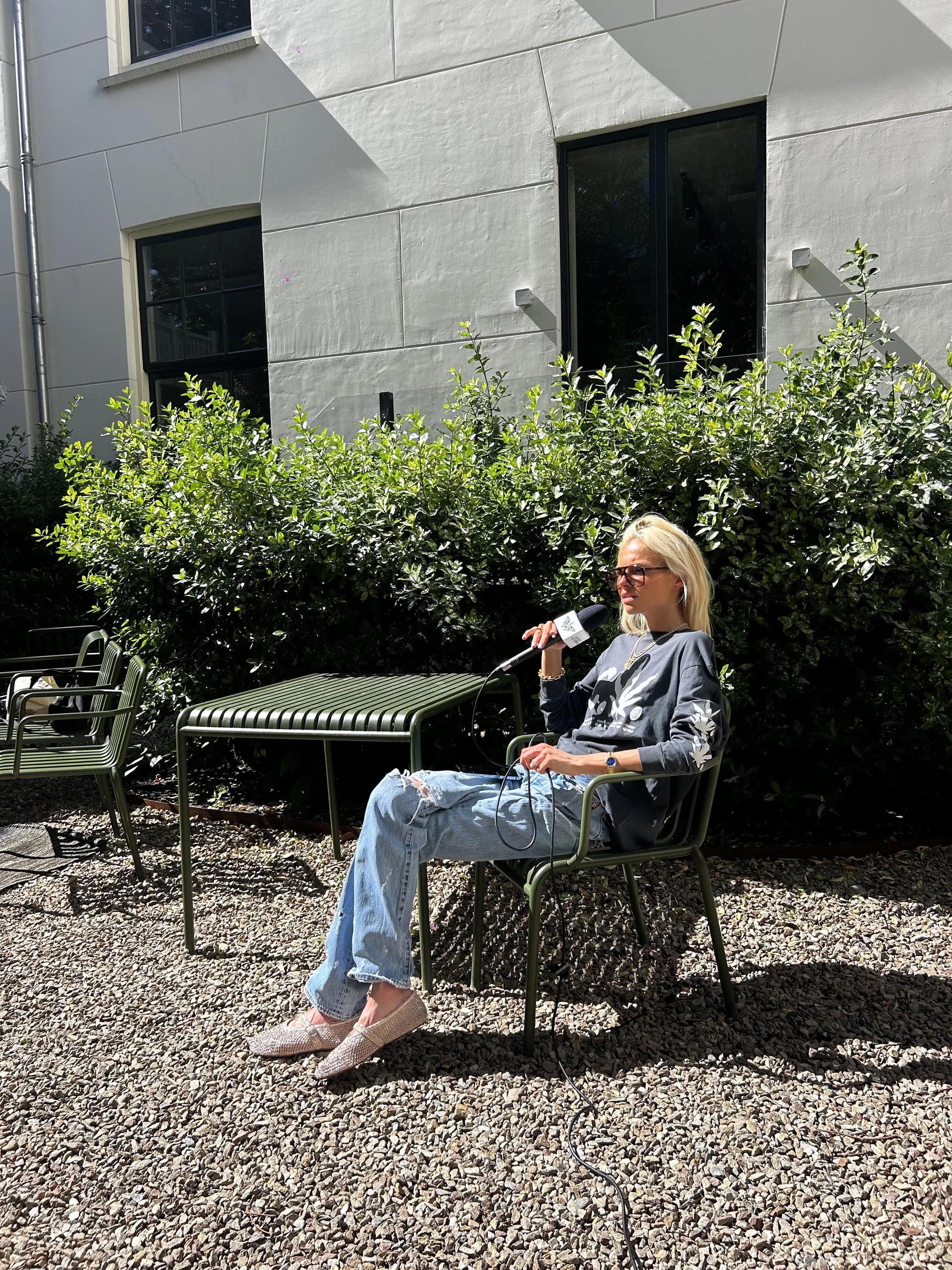 A woman with blonde hair, glasses, wearing a dark long-sleeve shirt with floral design, ripped jeans, and slip-on shoes, sitting outdoors on a green metal chair, holding a microphone with a logo, in front of a bushy hedge and a modern white building.