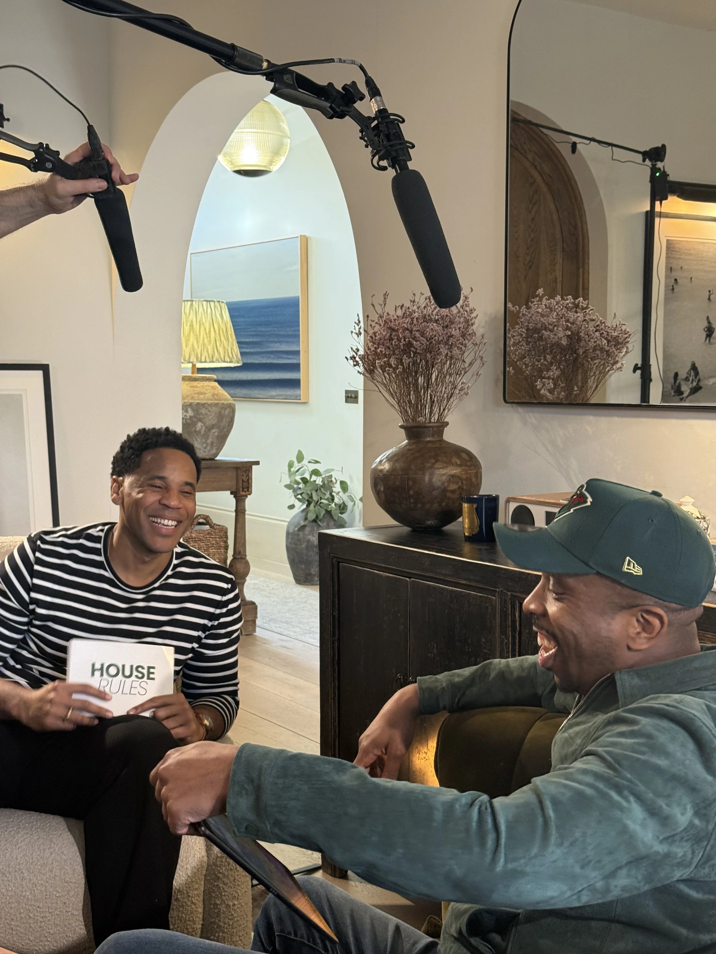 Two men sitting on the floor in a living room, holding hands and smiling. One man is wearing a striped shirt and the other is wearing a gray jacket and a green cap. A person is holding a boom microphone above them, indicating a recording or interview.