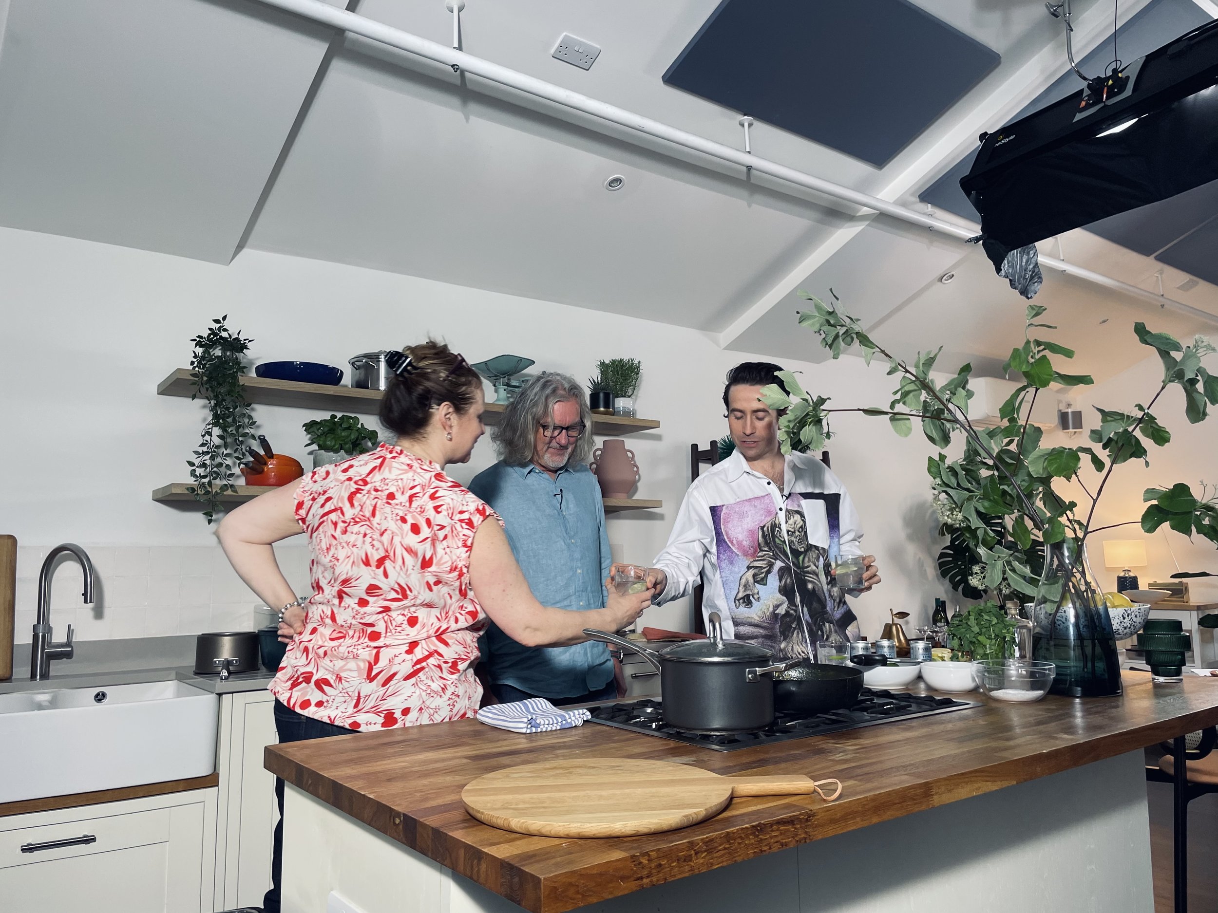Three people are gathered in a modern kitchen, engaged in conversation while preparing food. The kitchen features white cabinets, wooden shelves decorated with plants, and a wooden countertop with various cooking utensils and ingredients. The ceiling has visible pipes and ceiling speakers, and there is a large leafy plant on the right side of the image.