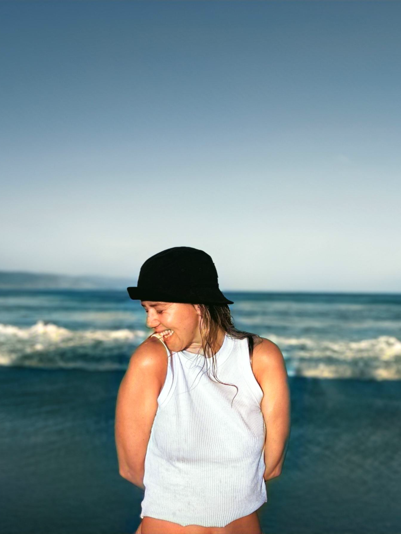 A woman wearing a black hat and white sleeveless top smiling on the beach with ocean waves and blue sky in the background.