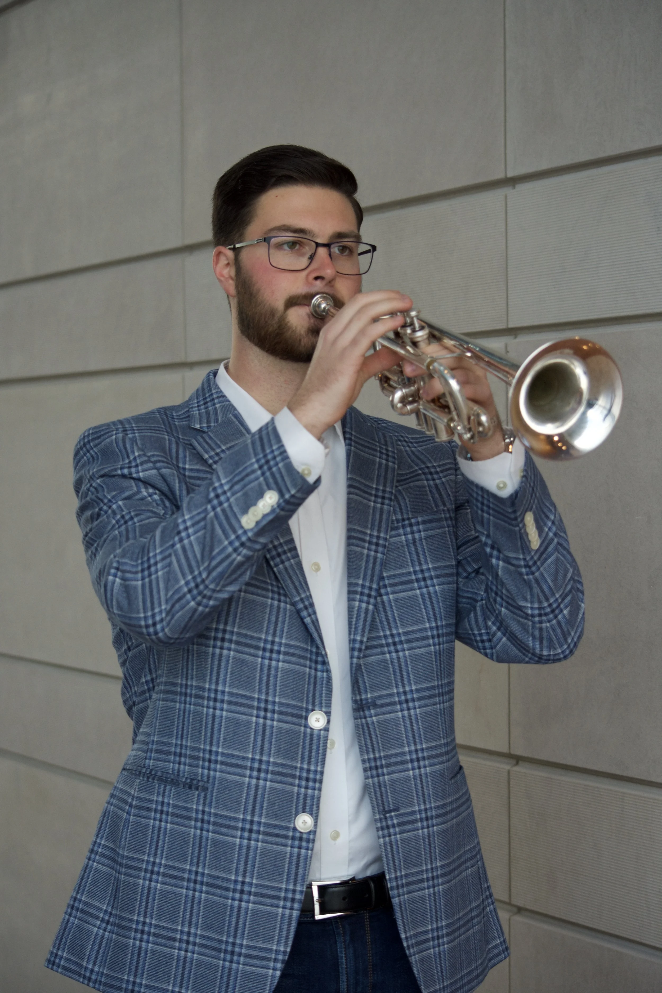 A man with glasses and a beard playing a trumpet, dressed in a blue plaid blazer, white shirt, and black belt, standing against a gray tiled wall.