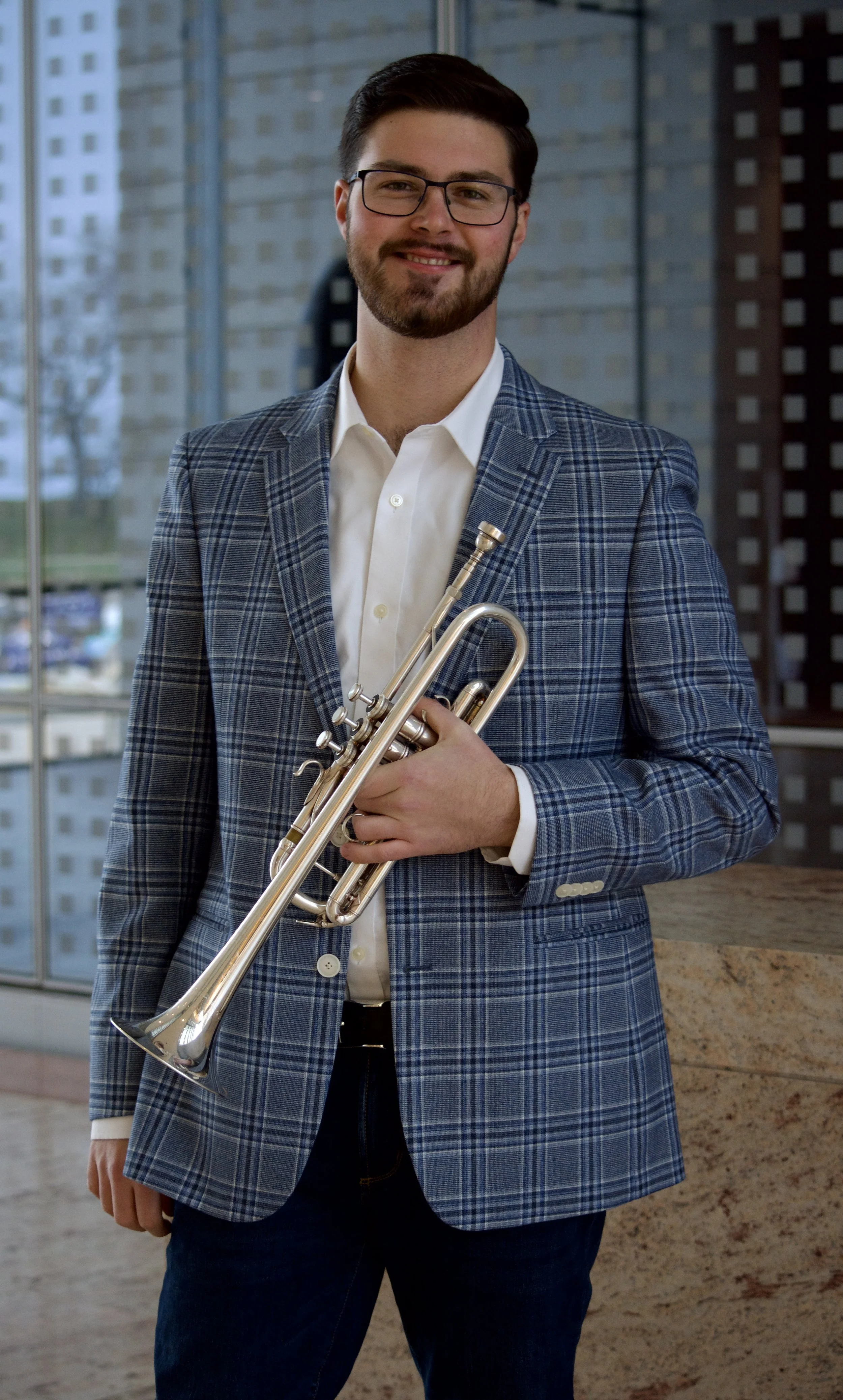 A man with glasses, a beard, and brown hair in a checkered blue blazer holding a trumpet in a modern indoor space with large windows.