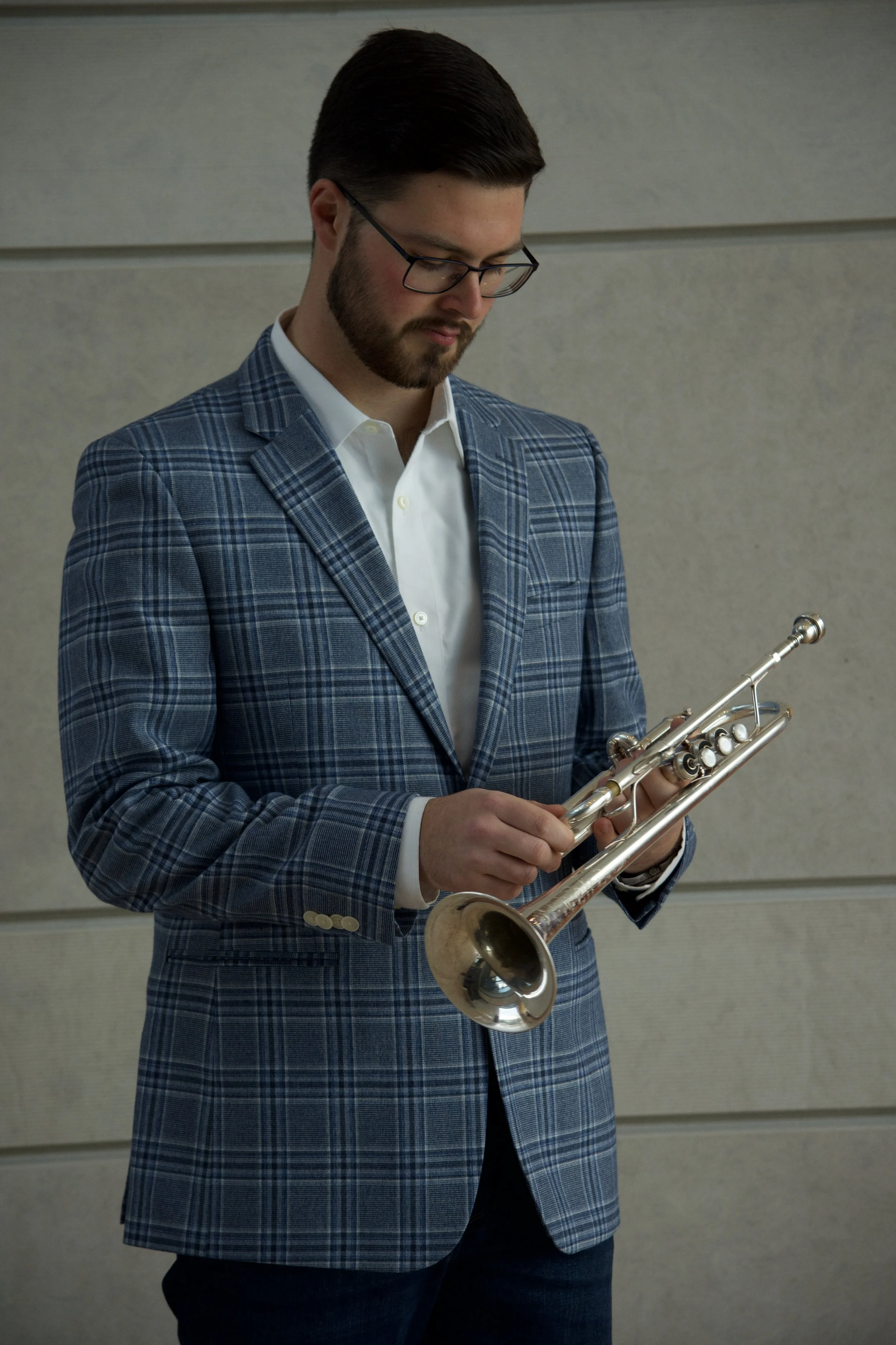 A man in a checkered blue blazer and white shirt holding a trumpet, standing against a plain, light-colored wall.