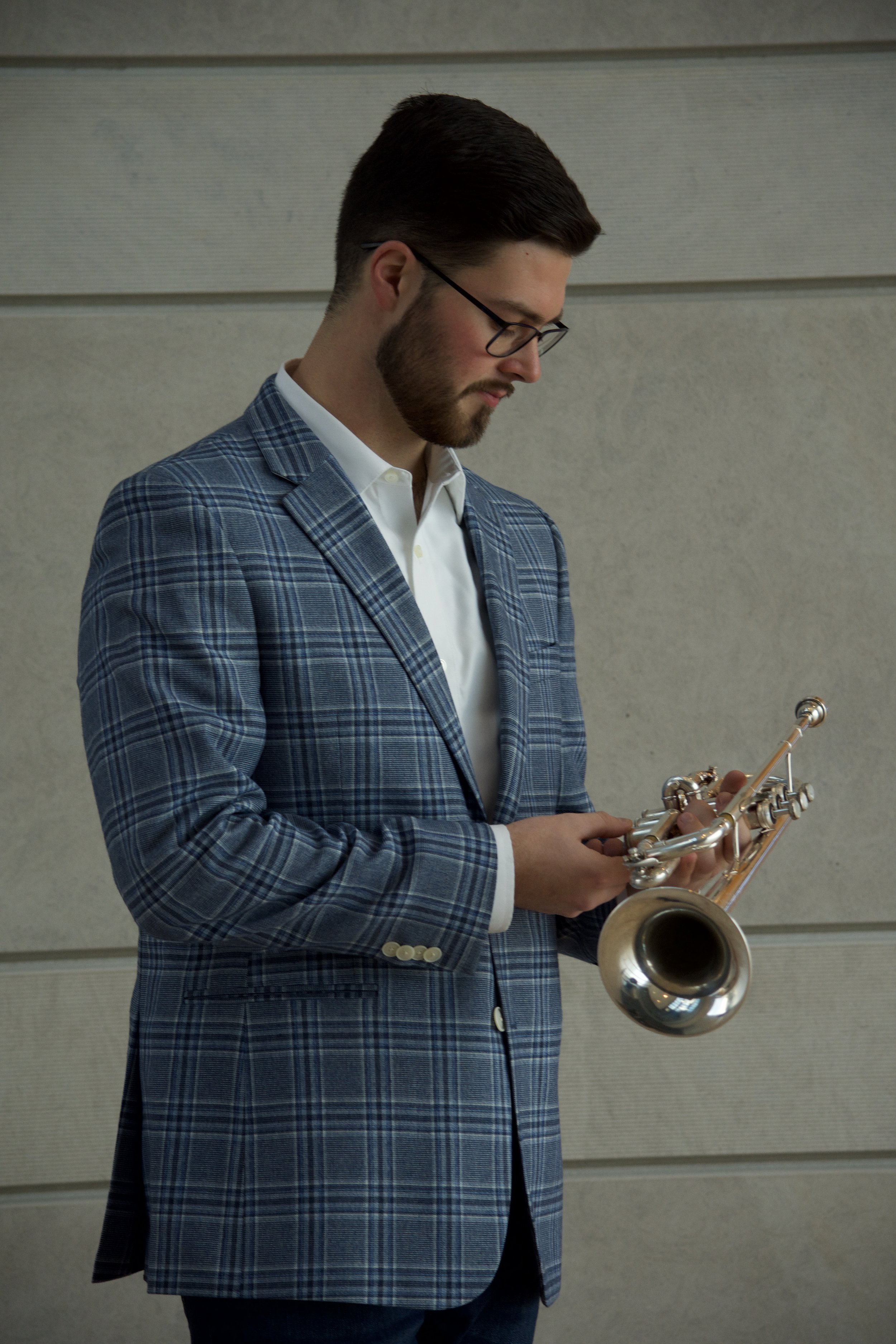 A man with dark brown hair, a beard, wearing glasses, a white shirt, and a blue plaid jacket, holding a trumpet and looking down at it against a beige wall background.