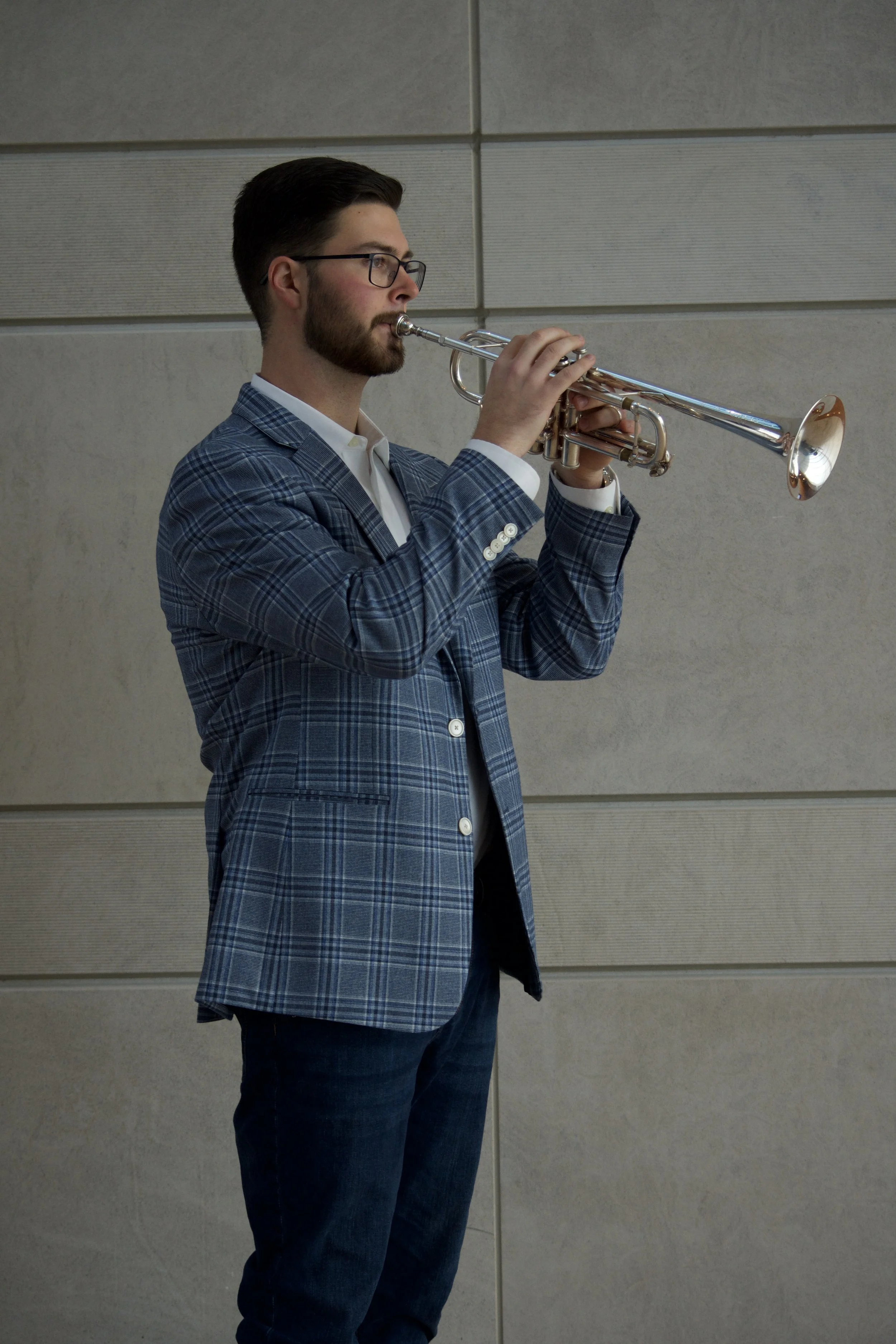 A man with glasses, dark hair, and a beard playing a silver trumpet. He is wearing a blue plaid blazer and a white shirt. The background consists of gray wall panels.