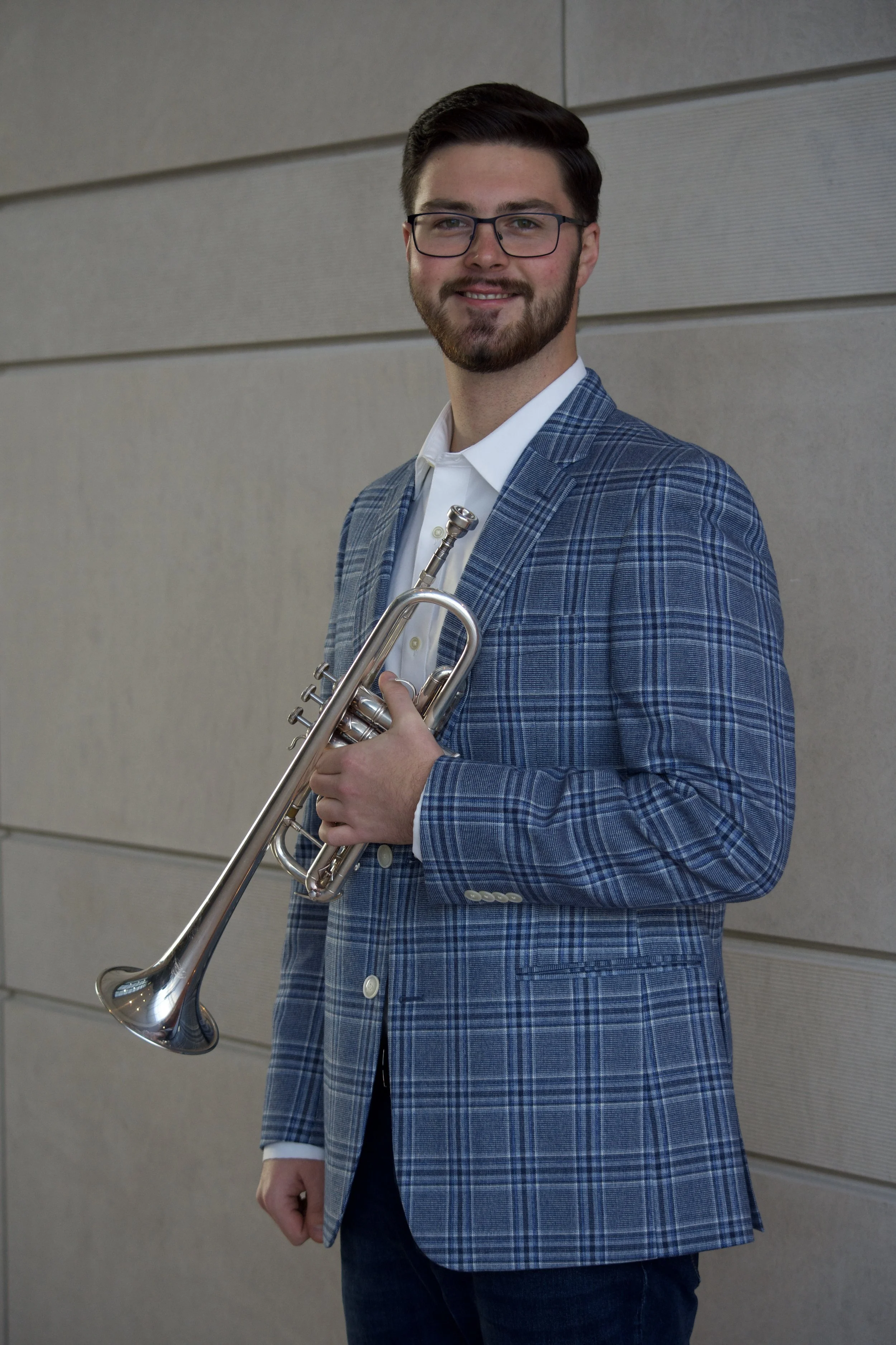 A man with dark hair, glasses, and a beard, dressed in a blue plaid blazer, white shirt, and dark pants, holding a trumpet, posing against a tiled wall.