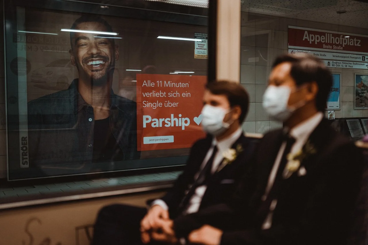Zwei Männer in formeller Kleidung und weißen Masken sitzen in einem U-Bahnhof, im Hintergrund ein Werbeplakat für eine Dating-App namens Parship, das einen lachenden Mann zeigt.
