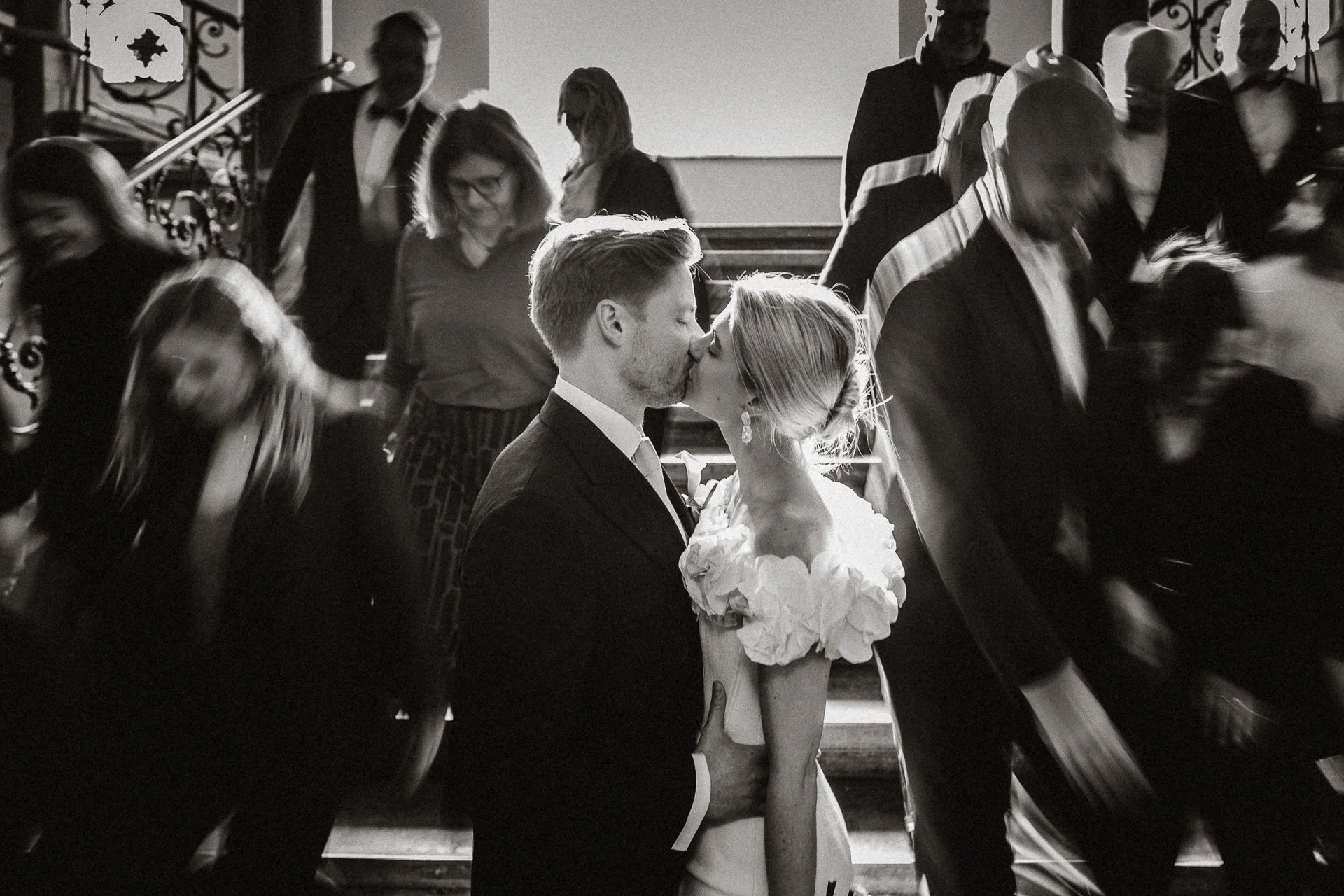 Ein Brautpaar küsst sich auf einer Treppe, umgeben von Gästen auf einer Hochzeit, schwarz-weiß Foto.