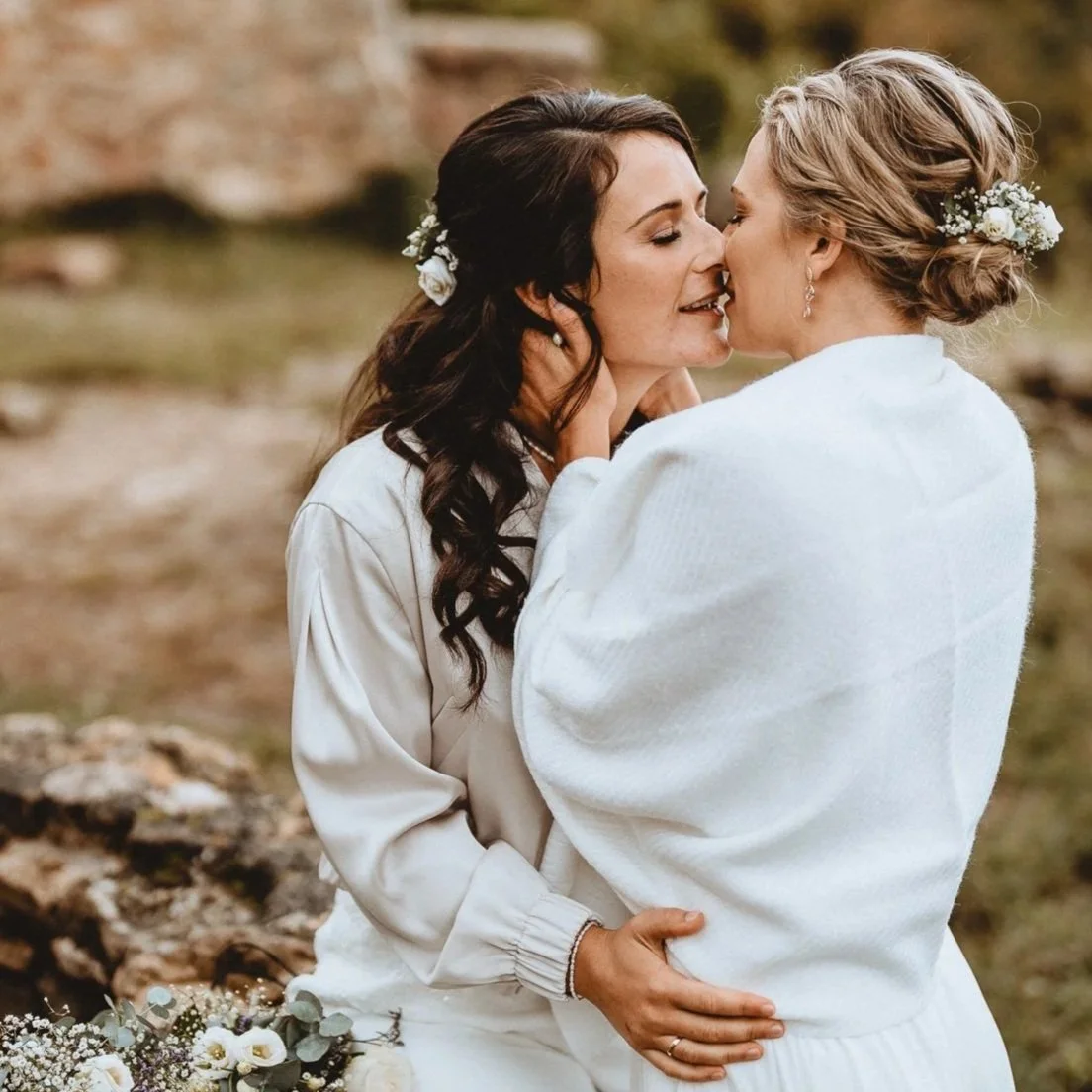 Zwei Frauen, die sich lieben und küssen, tragen weiße Kleidung und Blumen im Haar, im Freien.