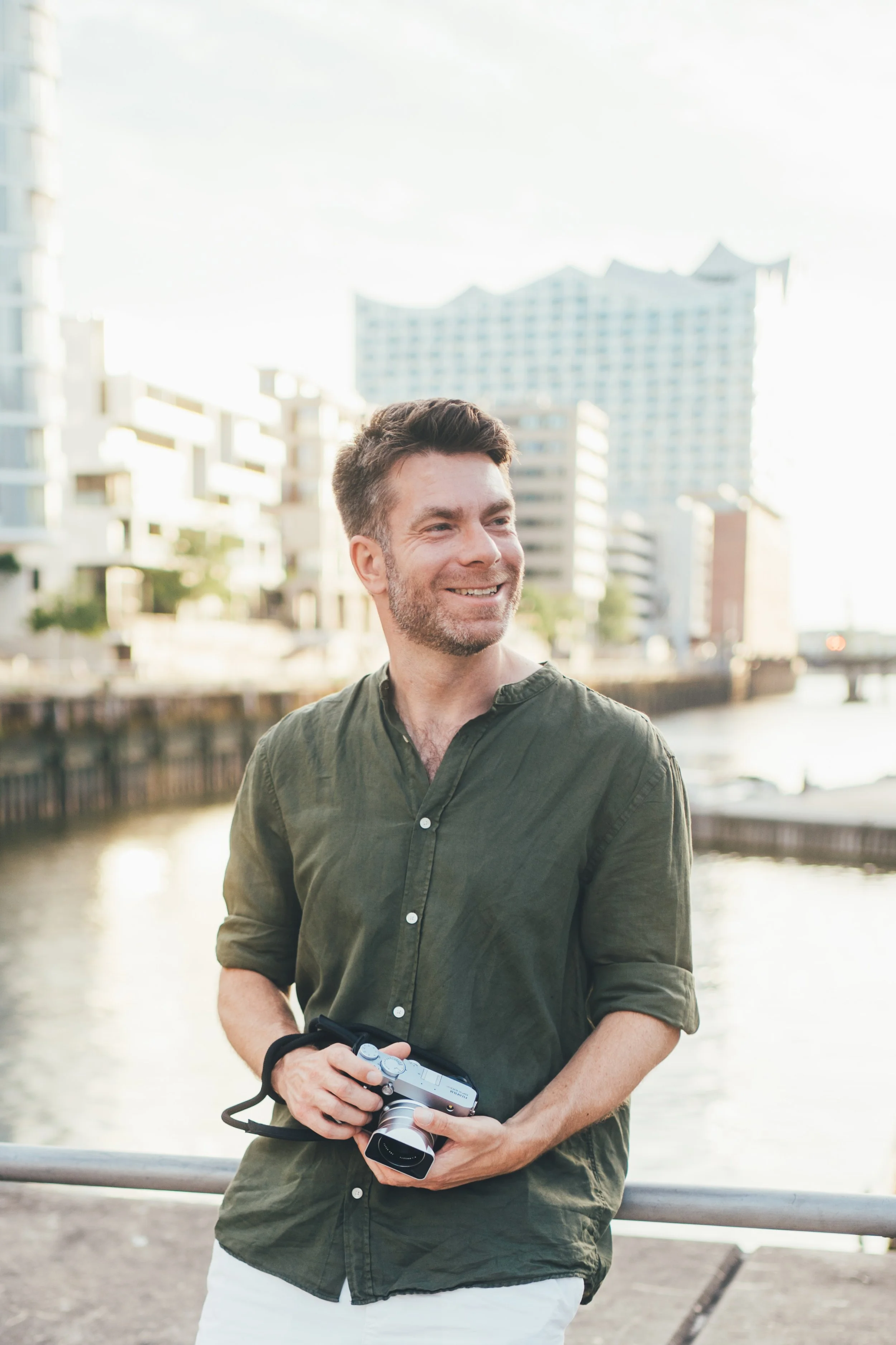 Ein junger Mann mit dunklem Haar und Bart, trägt ein grünes Hemd und hält eine Kamera in der Hand, steht vor einem Fluss in einer Stadt mit modernen Gebäuden im Hintergrund, bei Tageslicht.