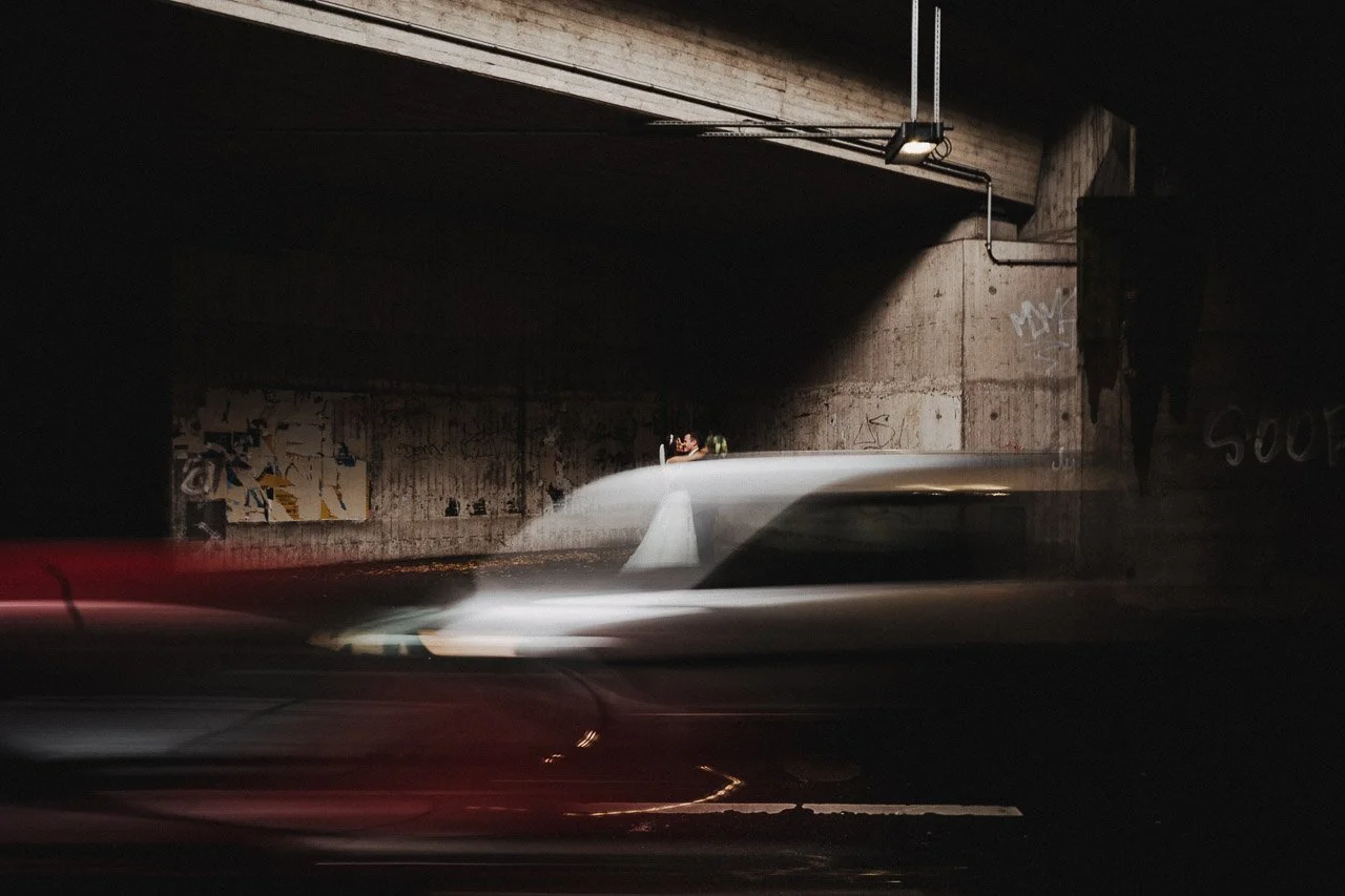 Ein Auto fährt an einer grauen Betonwand unter einer Brücke vorbei, bei Nacht, mit Bewegungsspuren und einem Graffiti an der Wand.