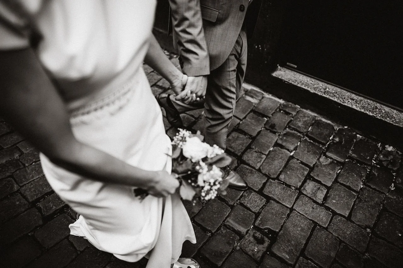 Ein Brautpaar hält sich an der Hand, die Frau trägt ein Hochzeitskleid und hält einen Blumenstrauß, sie stehen auf einer schmutzigen, gepflasterten Straße.