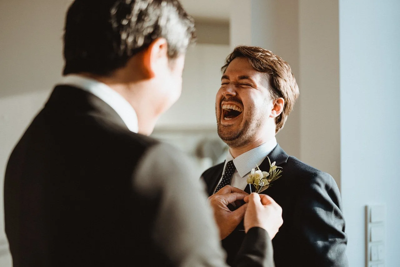 Zwei Männer in Anzügen lachen, während einer eine Blume an den Revers des anderen steckt.