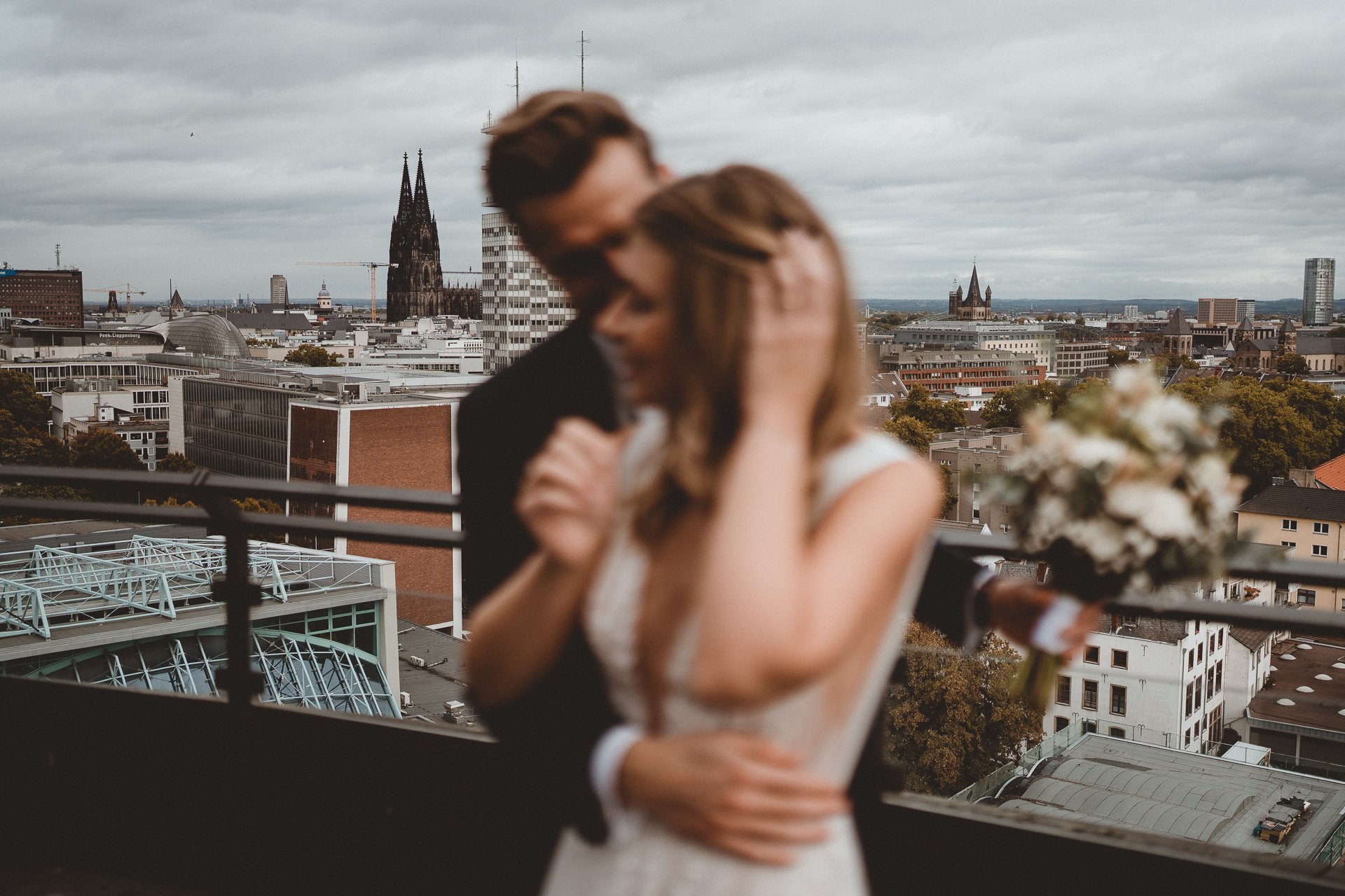 Ein verschwommenes Bild eines Paares, das sich auf einem Balkon mit Blick auf die Stadt Köln umarmt, im Vordergrund ein Blumenstrauß, mit Dom und weiteren Gebäuden im Hintergrund bei bewölktem Himmel.
