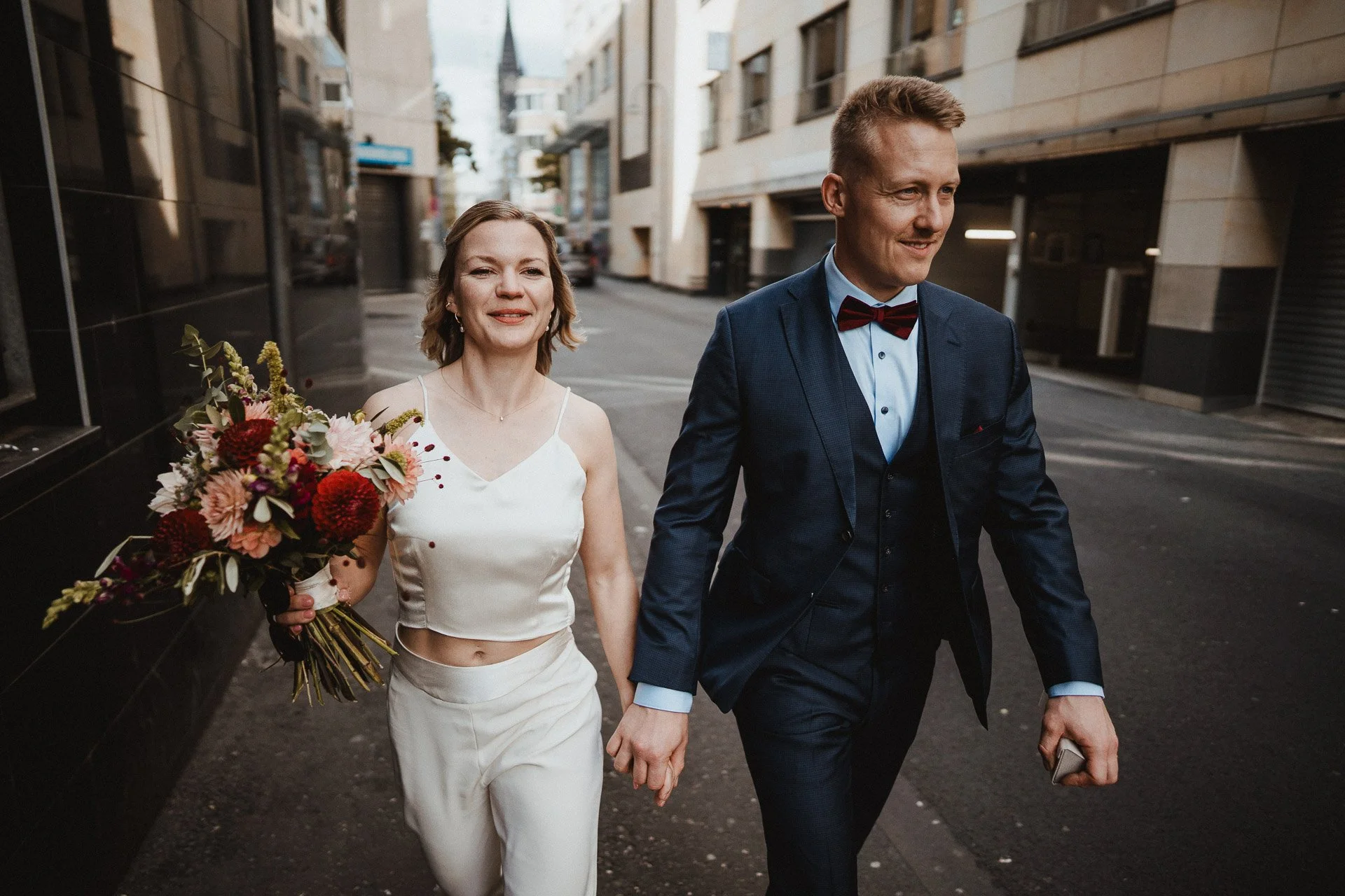 Ein Paar bei einer Hochzeit, die Frau trägt ein weißes Hochzeitskleid und hält einen Blumenstrauß, der Mann trägt einen dunklen Anzug mit Fliege, sie gehen gemeinsam auf einer Stadthstraße.