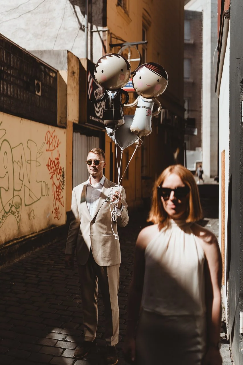 Ein Mann in Anzug hält drei Ballons, die ein Baby- und ein Hochzeitspaar darstellen, während eine Frau mit Sonnenbrille im Vordergrund steht.