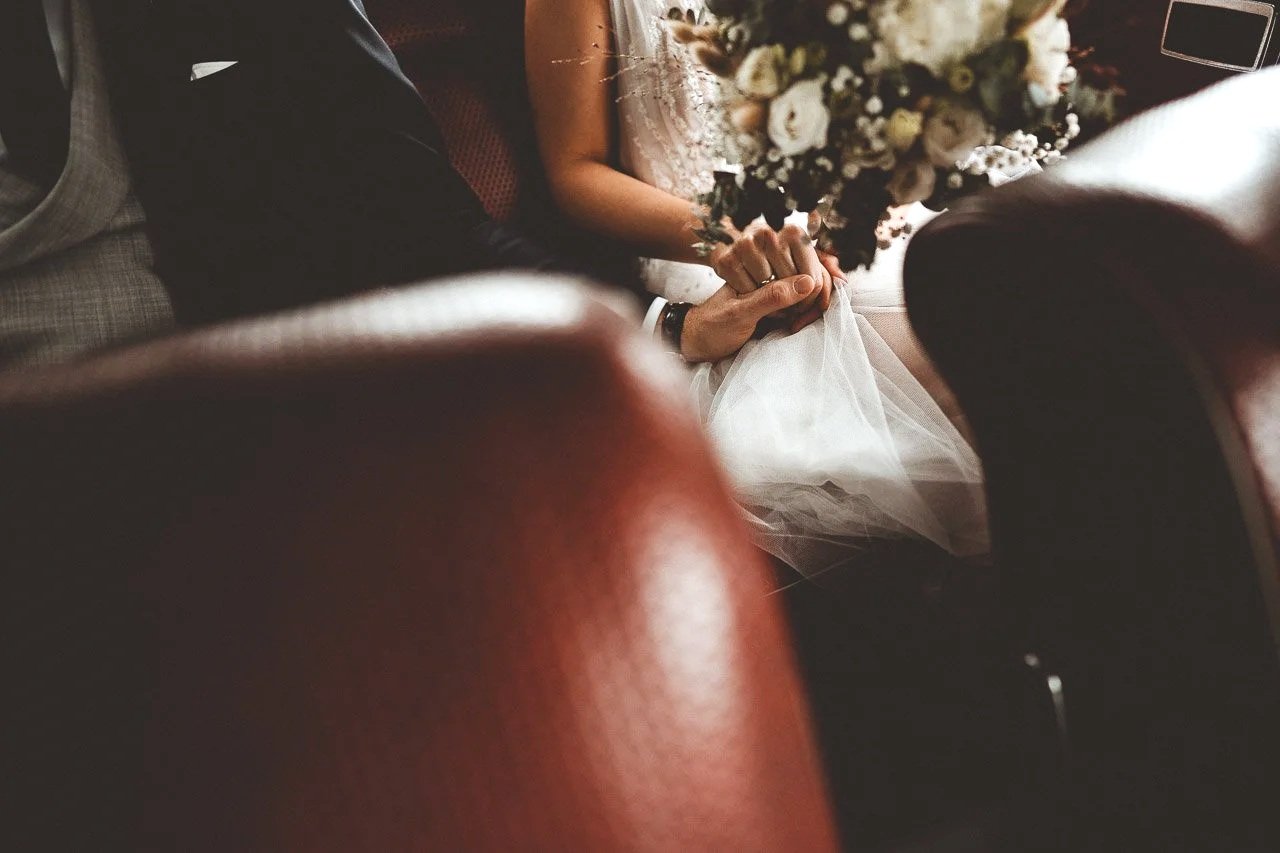 Ein Brautpaar sitzt auf einer Couch, die Hände sind verschränkt, im Vordergrund eine große Blumenbouquet.