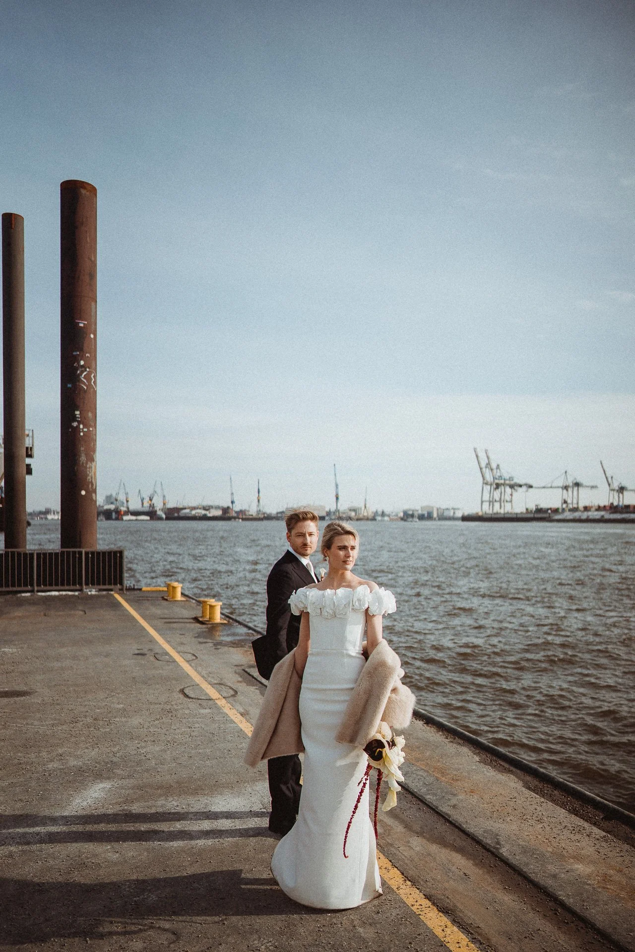 Ein Paar in formeller Kleidung steht an einem Hafen mit Wasser, Kräne und Schiffe im Hintergrund.