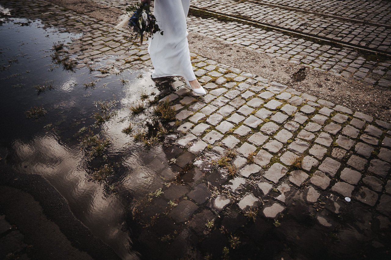 Eine Person in weißem Kleid und weißen Schuhen schreitet über eine nasse, gepflasterte Straße mit Pfützen und kleinen Pflanzen.