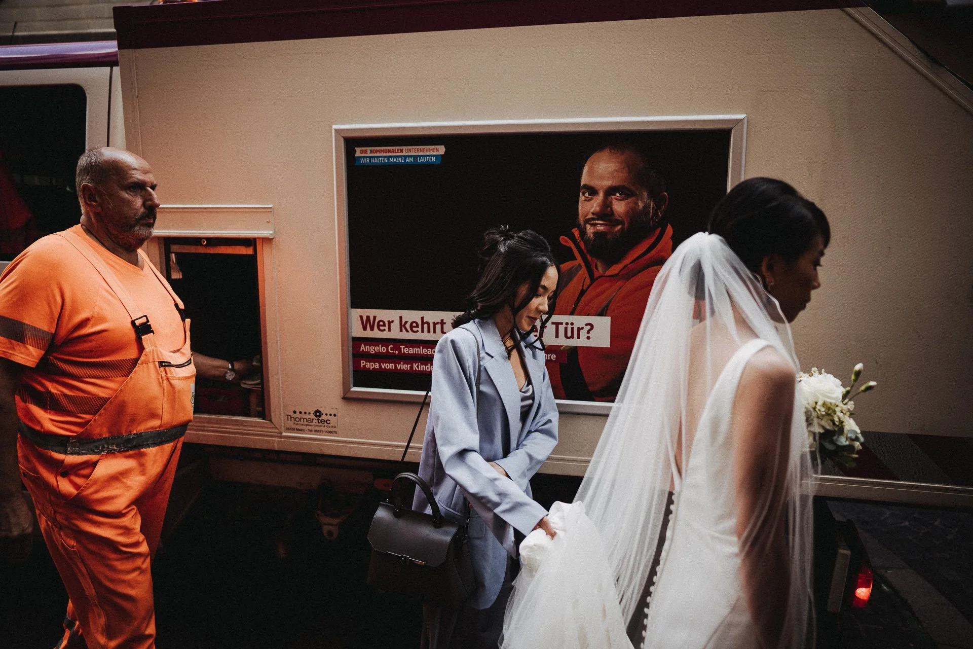 Ein Mann in orangener Weste und Frauen in Brautkleid stehen vor einem Poster, das eine männliche Person zeigt. Es ist eine Szene, die vermutlich in einem öffentlichen Gebäude stattfindet.
