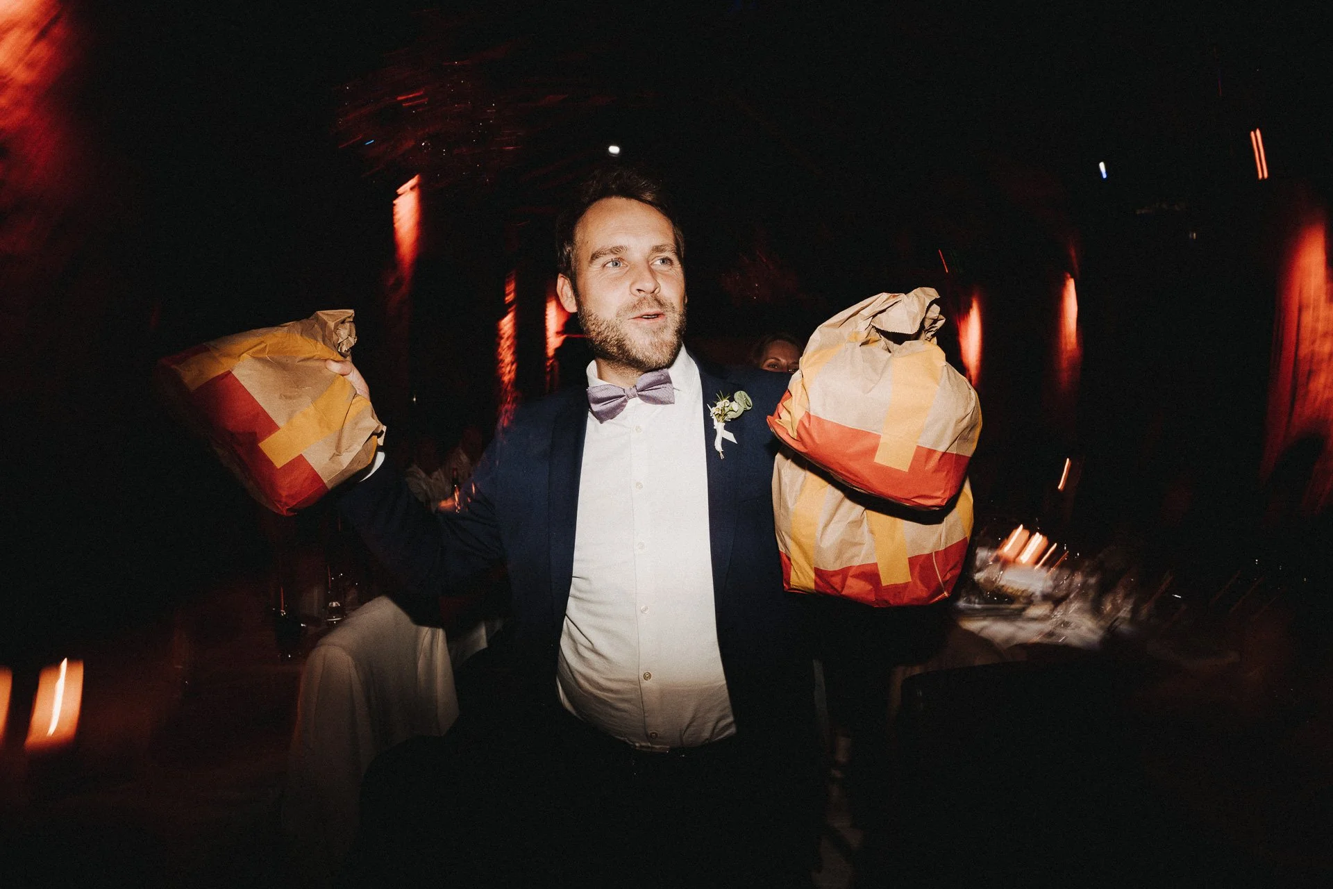 Ein Mann in Hemd, Anzug und Fliege trägt zwei große Papiertüten auf den Schultern auf einer Party mit dunklem Hintergrund und roter Beleuchtung.