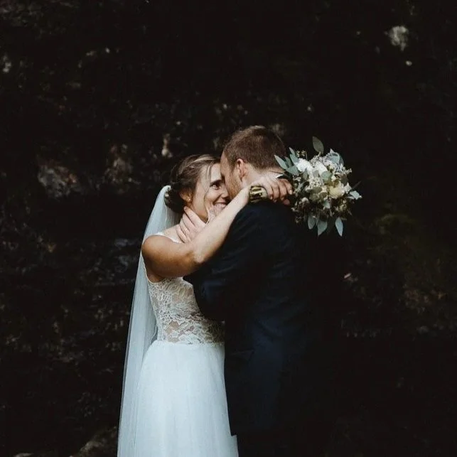 Braut und Bräutigam küssen sich bei ihrer Hochzeit, sie umarmt ihn und hält einen Brautstrauß, im Hintergrund ist dunkler Naturhintergrund.