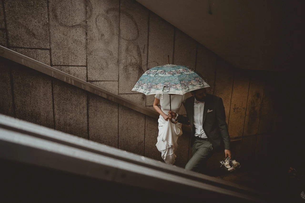Ein Mann und eine Frau gehen die Treppe in einem Gebäude hinauf, die Frau hält einen Regenschirm und trägt ein weißes Kleid, der Mann trägt einen schwarzen Anzug und hält Blumen.