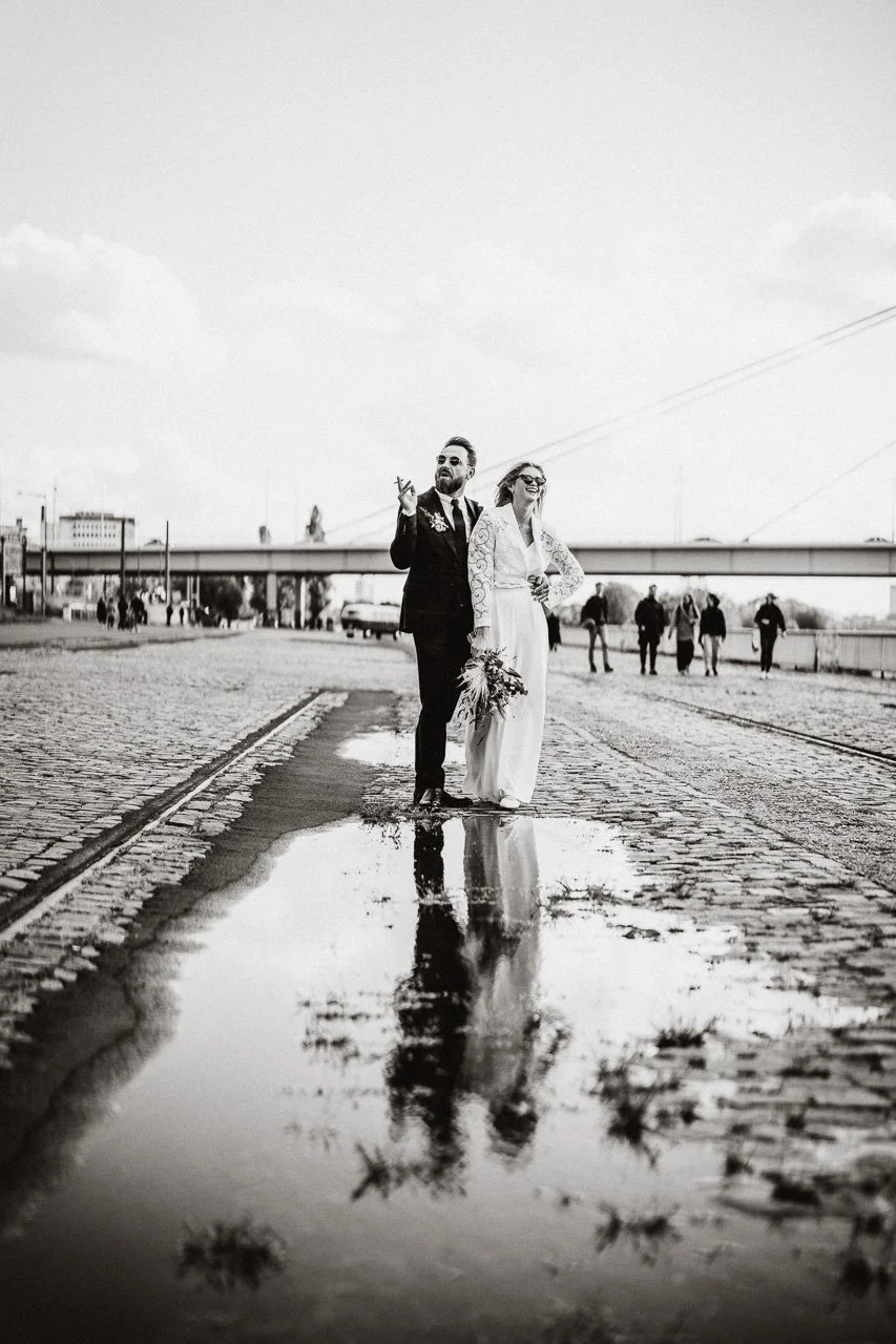 Ein Brautpaar in Hochzeitskleidung, das auf einer regennassen gepflasterten Straße steht, mit spiegelnder Wasserpfütze im Vordergrund, im Hintergrund einige Passanten und eine Brücke, alles in Schwarz-Weiß.