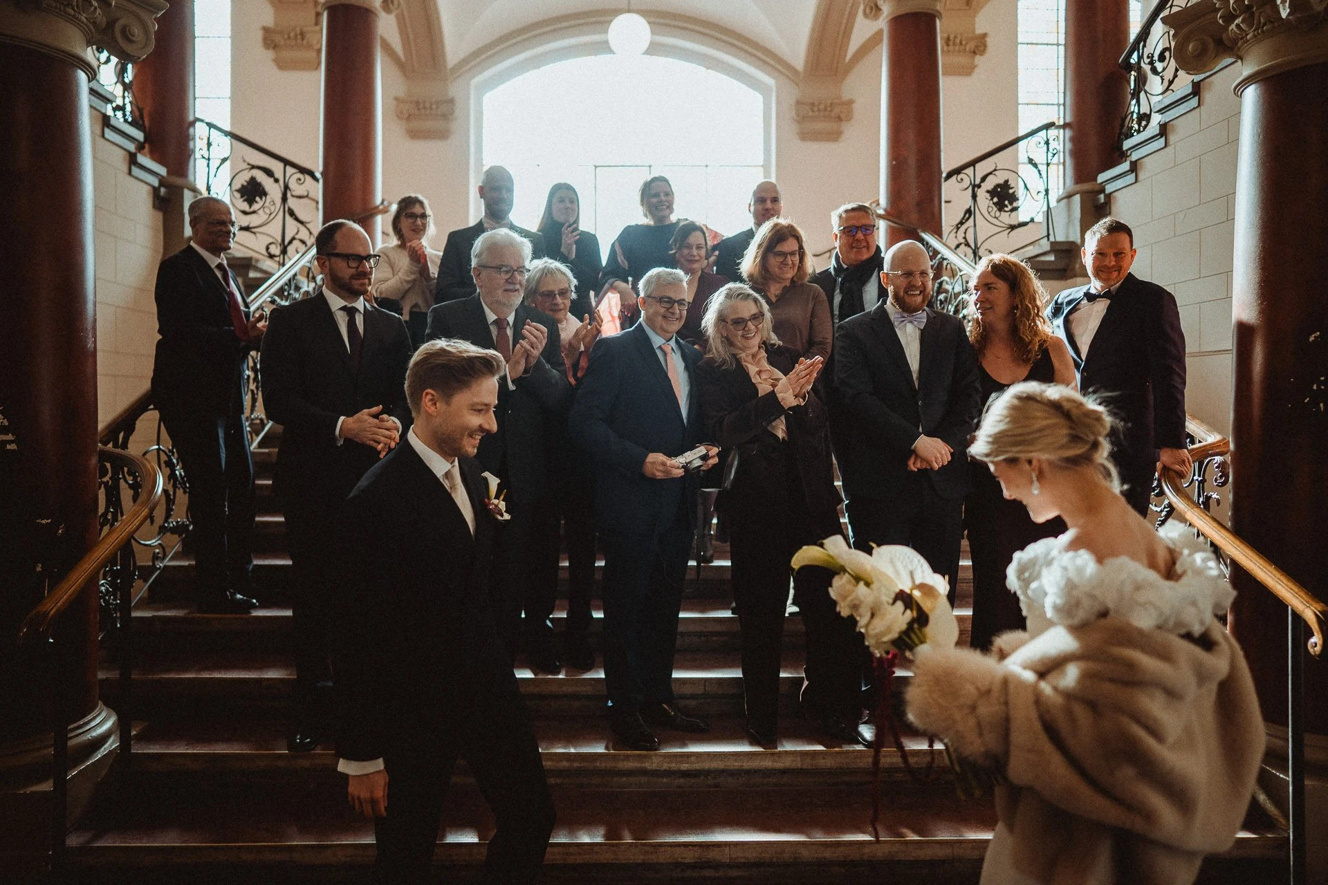 Hochzeitsgesellschaft versammelt sich auf einer Treppe in einem eleganten, historischen Gebäude, die Braut mit Brautkleid hält Blumengebinde in der Hand, während die Gäste lächeln und applaudieren.