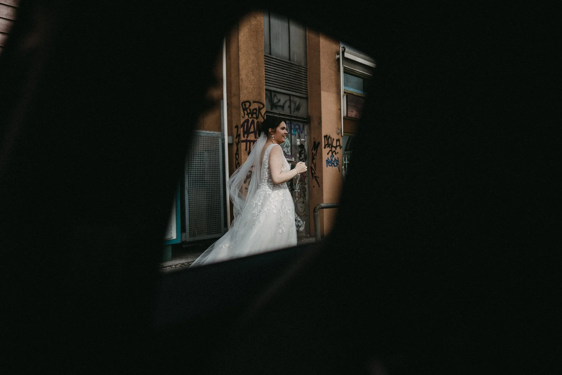 Braut in Hochzeitskleid auf einer urbanen Straße, umgeben von Graffiti, durch eine dunkle, unregelmäßig geformte Lücke sichtbar.