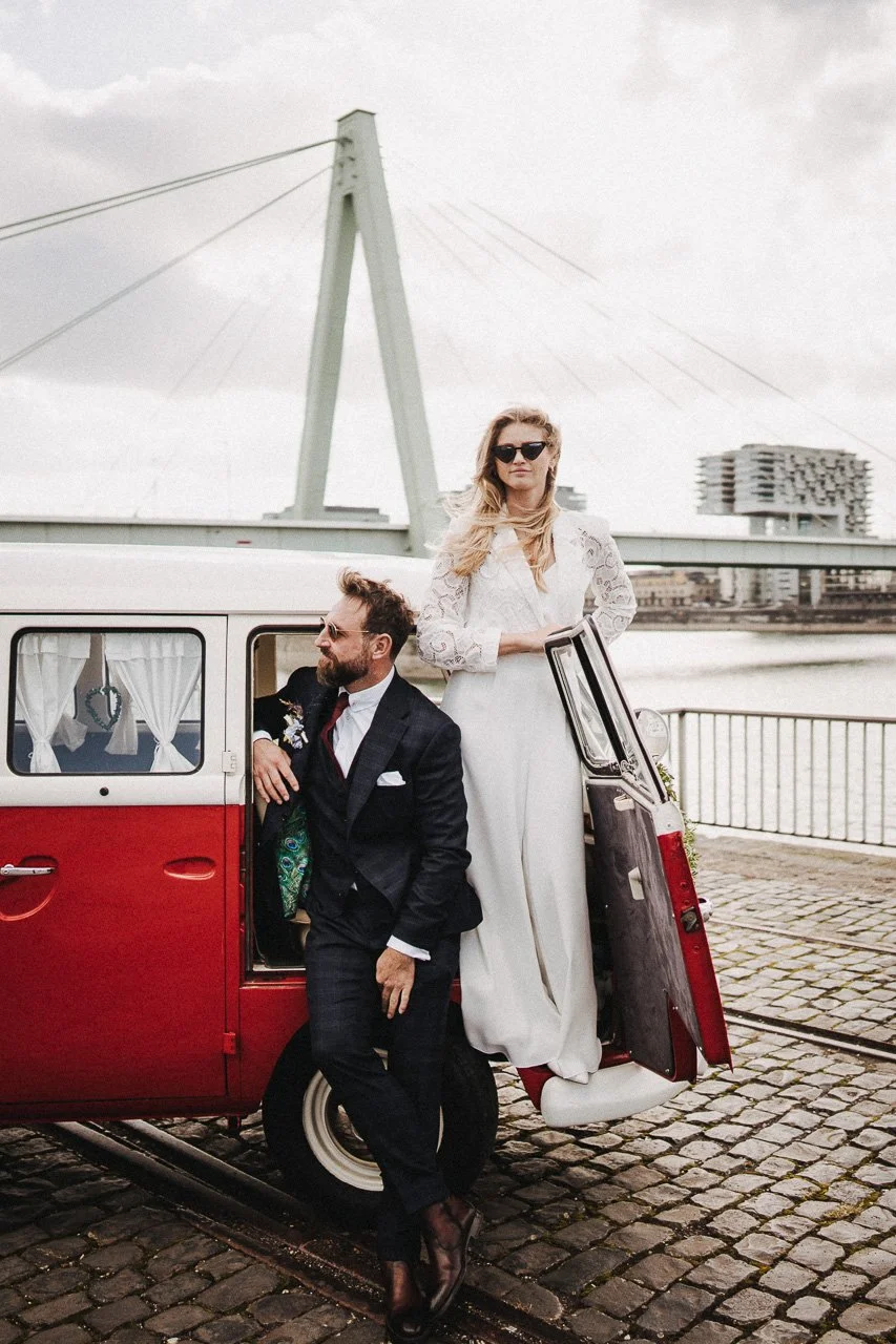 Ein Mann in einem schwarzen Anzug und eine Frau in einem weißen Kleid stehen neben einem roten Oldtimer an einer Uferpromenade, im Hintergrund eine Brücke und ein modernes Gebäude.