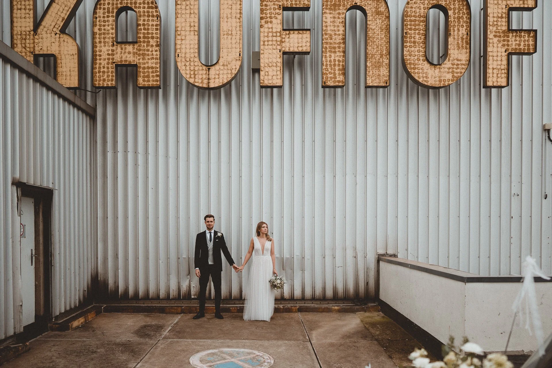 Ein frisch verheiratetes Paar hält Händchen, vor einer großen Wand mit Metallbuchstaben, die vermutlich das Wort 'HOPE' oder 'LOVE' bilden, in einer urbanen Umgebung.