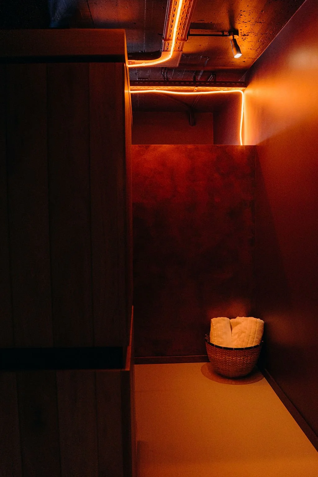 A dimly lit room with dark wood paneling, orange ambient lighting along the ceiling and walls, and a basket with towels in the corner.