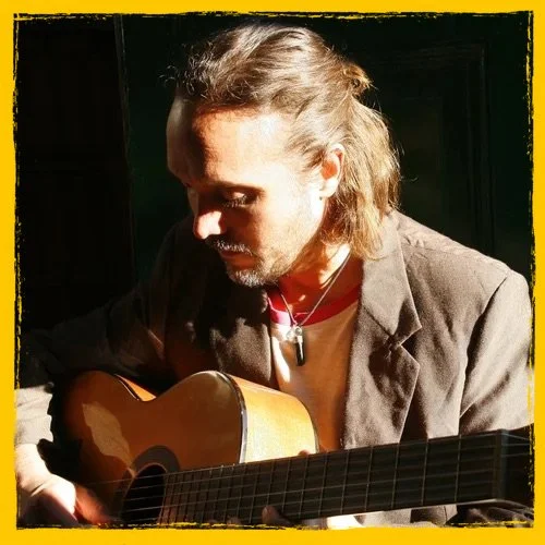 A man with long hair and a beard playing an acoustic guitar indoors, with warm lighting and a dark background.