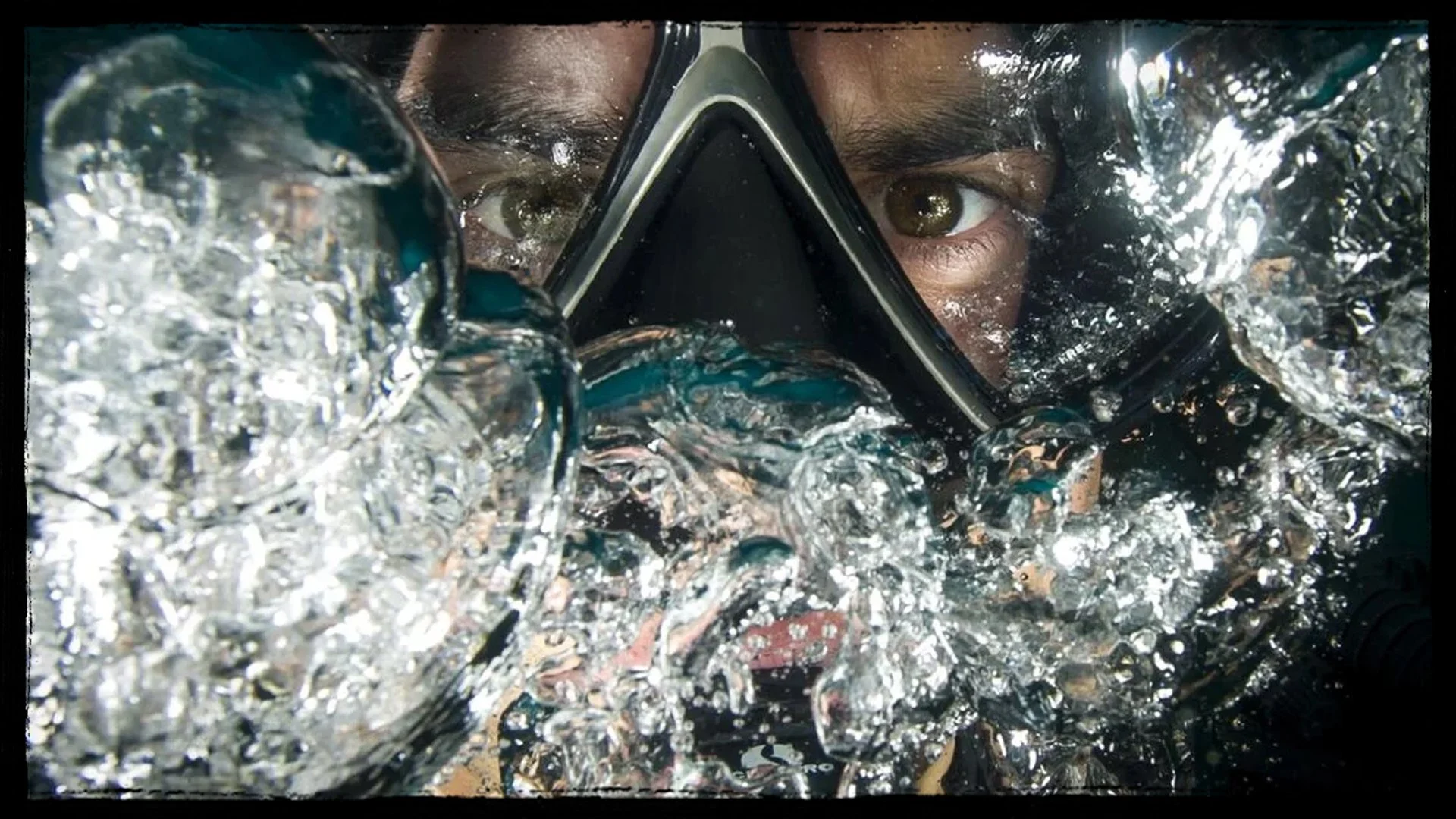 A man in snorkeling gear with a mask and snorkel surrounded by splashing water, looking directly into the camera.