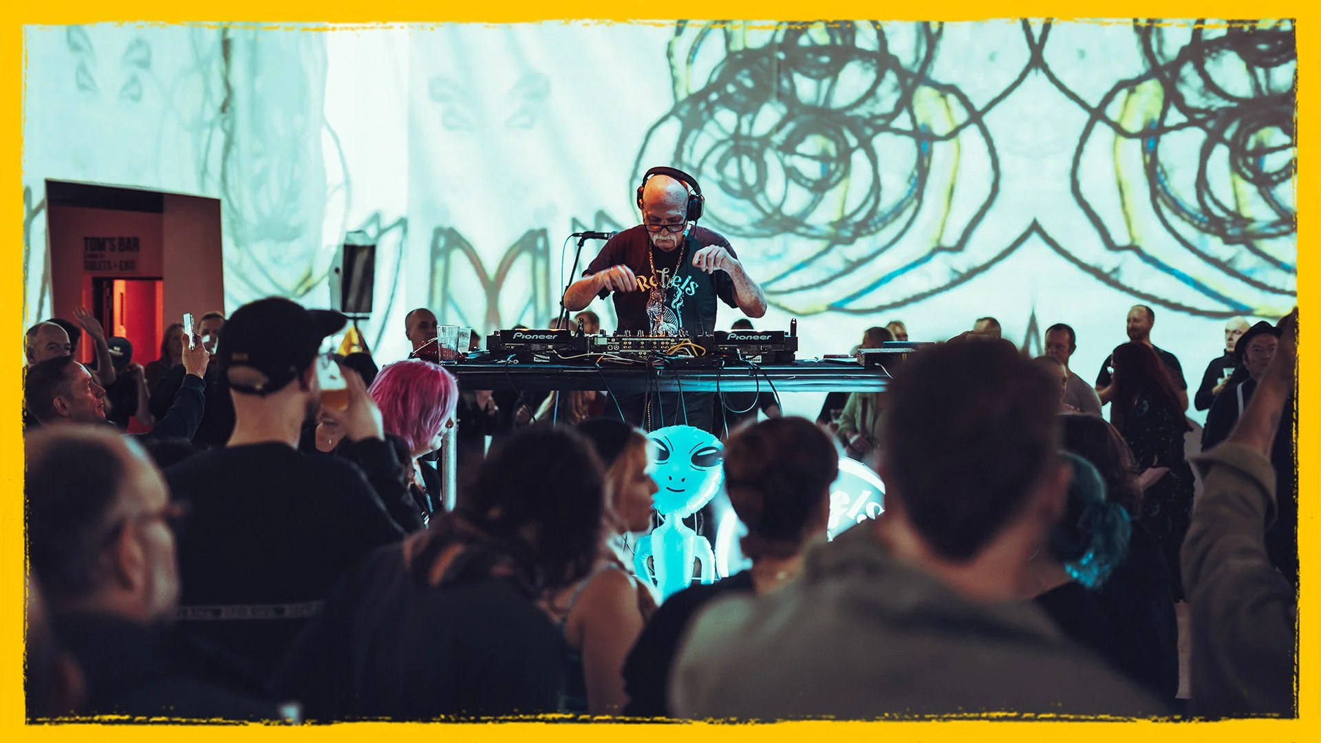 A DJ performs on stage with a crowd dancing and watching. The background features abstract art projected on a large screen.