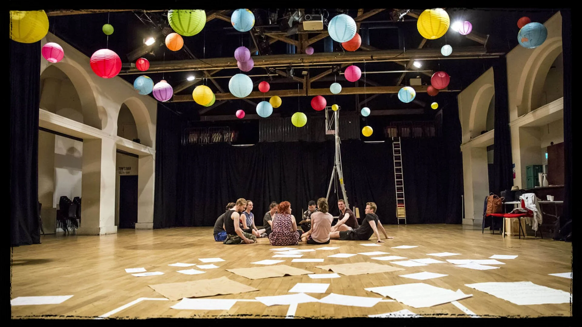 People sitting in a circle on a wooden floor in a theater with colorful hanging lanterns overhead and papers scattered on the floor.