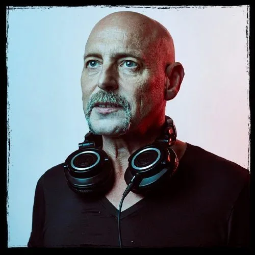 A man with a bald head, mustache, and goatee wearing a black shirt and black headphones around his neck, against a gradient background.