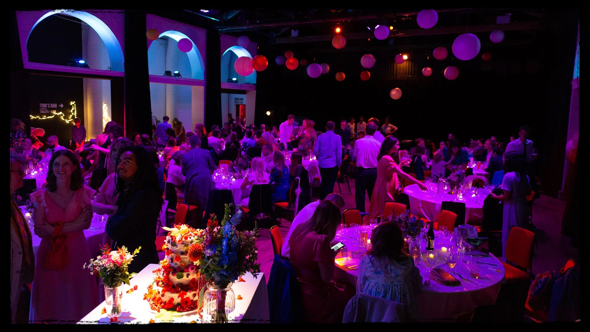 A lively, colorful party or celebration in a dimly lit venue with purple lighting and hanging paper lanterns, filled with numerous round tables of people chatting, dining, and enjoying the event.