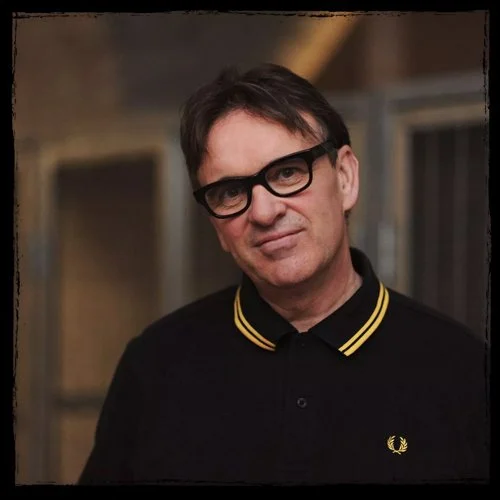 A man with dark hair, wearing black-rimmed glasses and a black shirt with yellow trim on the collar, standing indoors.
