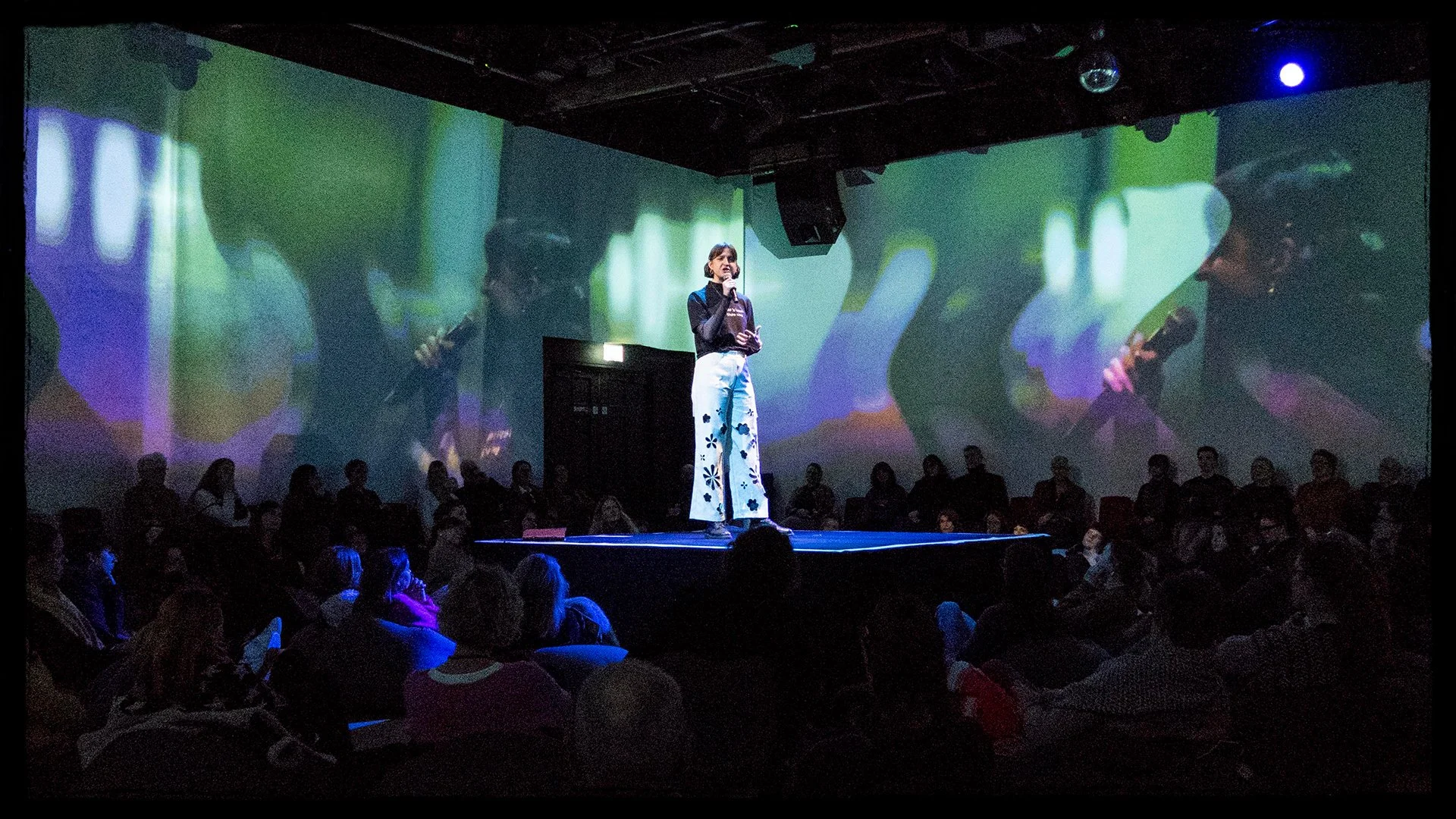 A woman stands on a stage with a microphone, speaking in front of an audience. The background features projections of a police officer with a microphone, with some of the audience members watching attentively.