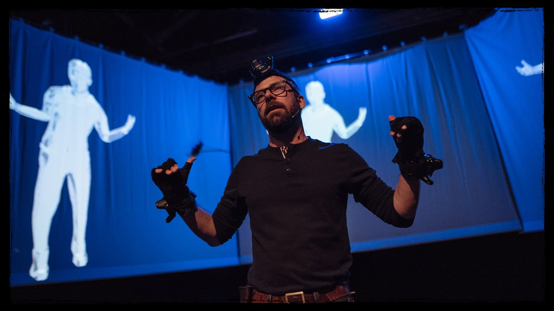 A man with a beard, glasses, and wearing black gloves and a black shirt stands on a stage with blue curtains and two large projected images of a person in white behind him. The man is gesturing with his hands and looking upward.